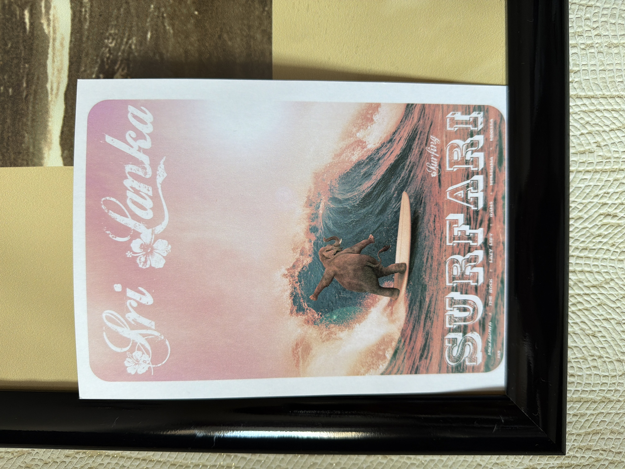 A framed advertisement for "Surfari" in Sri Lanka. It features an elephant surfing on a wave. The frame is black and the poster is white with pink and brown accents. The advertisement also includes the words "Sri Lanka".