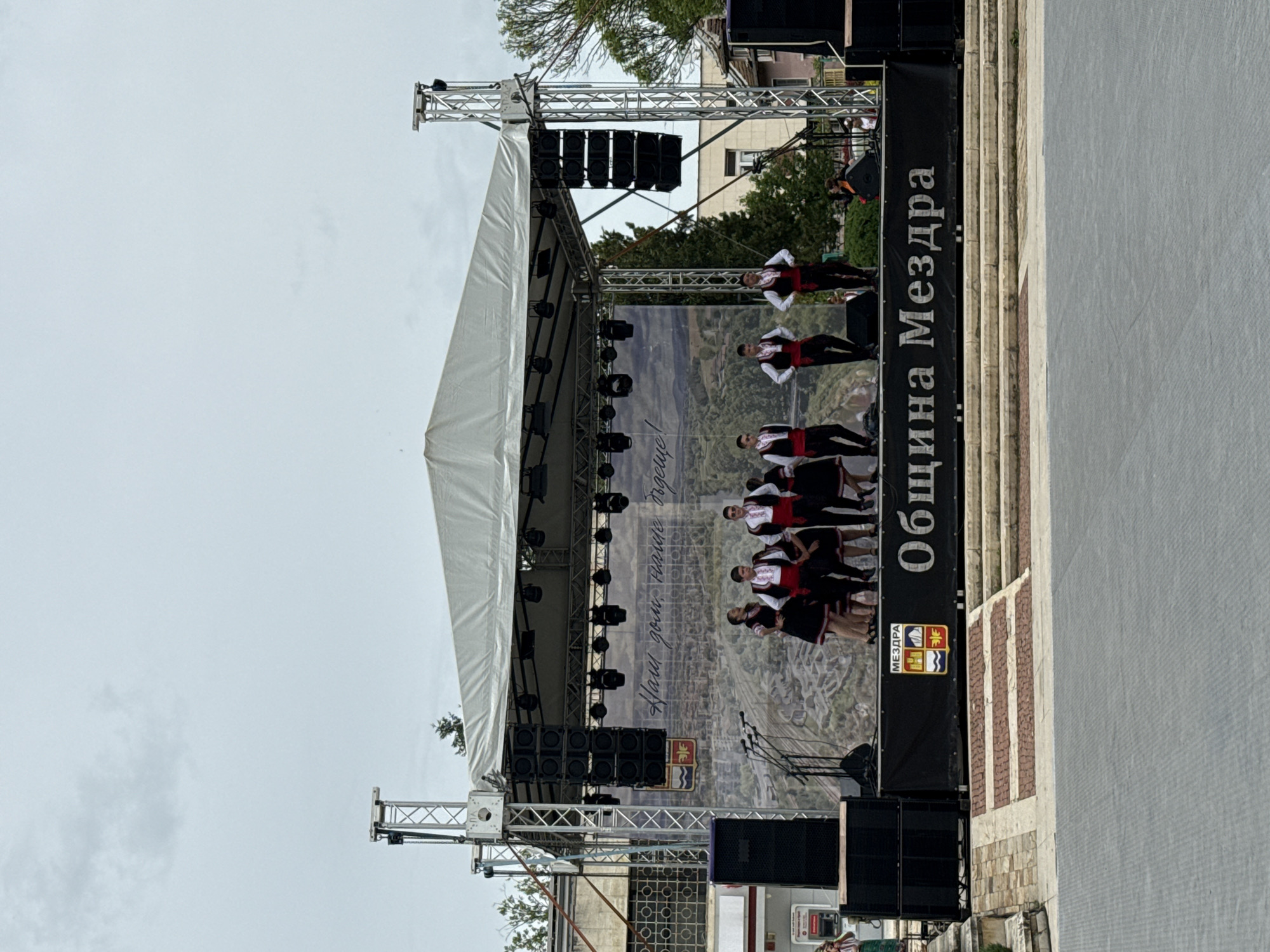 A stage is set in the city of Mezdra, Bulgaria. People dressed in traditional Bulgarian folk costumes are performing on stage. Banners reading "Община Мездра" and "Наш дом! Наше бъдеще!" are visible, along with the coat of arms. The stage is equipped with lighting and sound equipment. The background shows an image of the city's landscape.