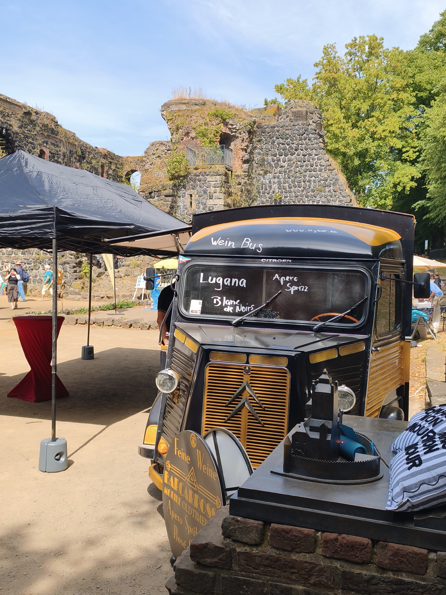 The image shows a black and yellow vintage Citroën van, repurposed as a "Wein Bus" (wine bus), parked outdoors. The van is adorned with text on its windshield and side, including "Wein Bus," "Lugana," "Apero," "Spri22," "Blanc de Noirs," and "www.hybar.de." A cylindrical sign with "Feine Weine" and "BARCAROSSO" is visible near the van's front. The van is set against a backdrop of ancient stone ruins and lush green trees under a clear blue sky. In the foreground, a black tent provides shade over a red tablecloth-draped table. Several people are visible in the background, walking and interacting. A model of a building is placed on a table in front of the van. The overall scene suggests an outdoor event or market, possibly a historical site given the ruins. The text on the van and sign indicates it is serving wine and aperitifs. The location is identified as the Ratingen Castle ruins in Ratingen, which is near Düsseldorf, Germany.