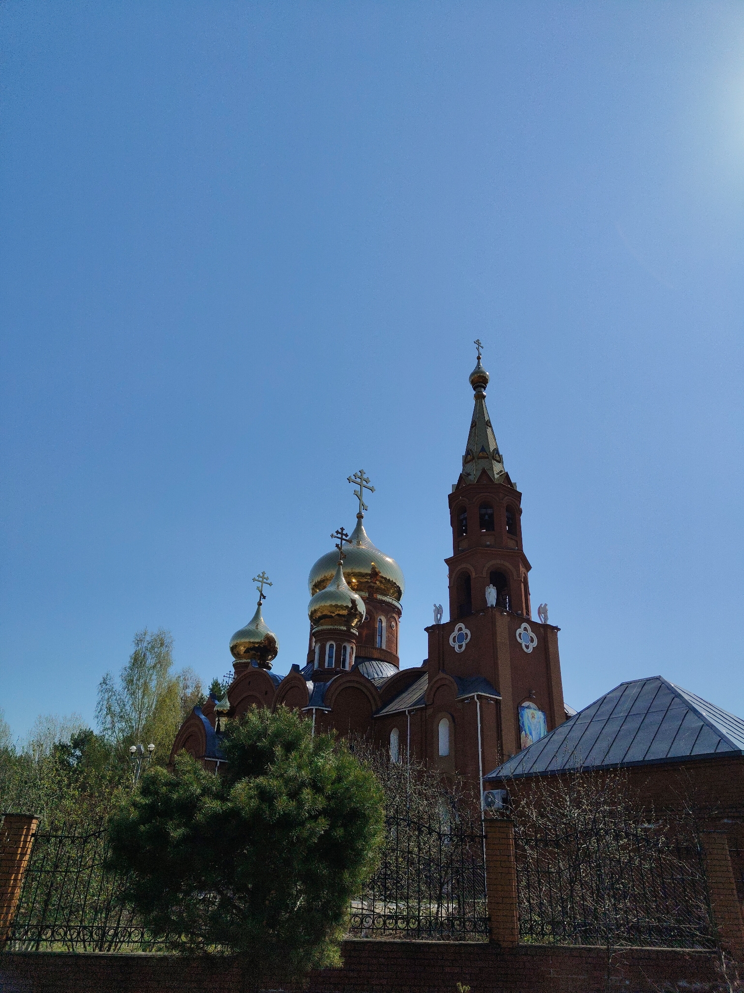 The image shows a brick Orthodox church located in an urban area of Unknown, Unknown, under a clear blue sky. The church features a red brick facade with gold domes and crosses. The primary dome is flanked by smaller ones, and a tall bell tower is visible to the right, adorned with statues. A brick wall with a black wrought-iron fence surrounds the church grounds, with a lush evergreen tree partially obscuring the view of the church from the front. To the side of the church, a low building with a gray roof is present. The background includes some deciduous trees with budding leaves, suggesting springtime. The lighting indicates a bright, sunny day.