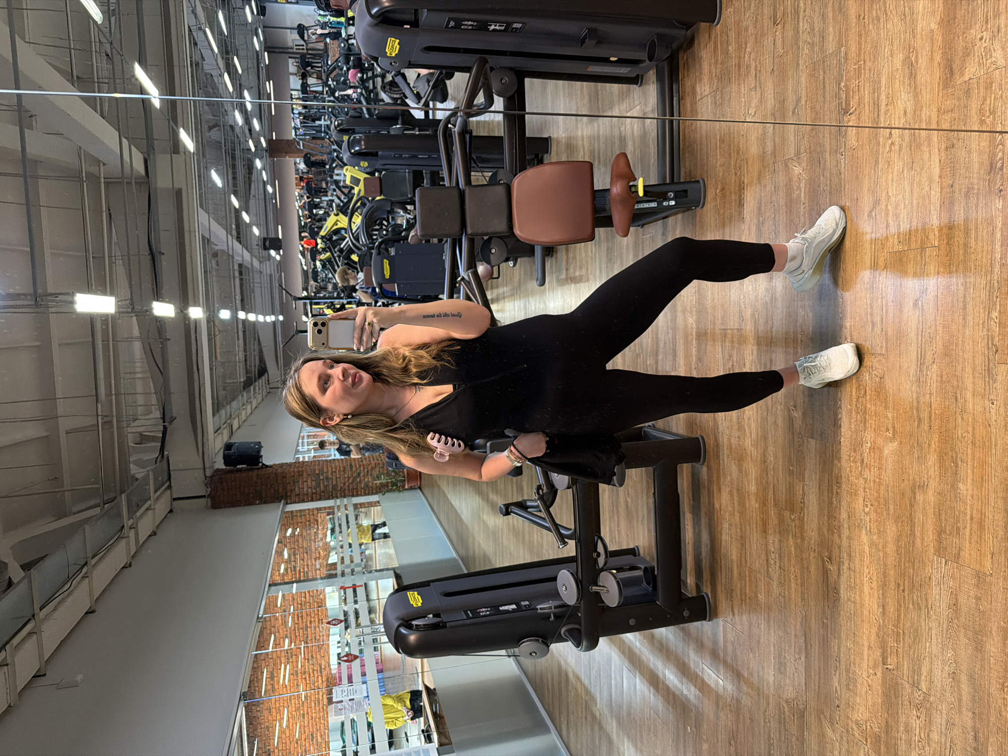 A young woman is posing for a selfie in a gym, likely located in Chaikovsky, Russia, as reflected in the mirrored wall showing external signage. She is wearing a black jumpsuit and white sneakers. The gym is well-equipped with various exercise machines, including leg press and cable machines. The interior features a wooden floor and exposed brick elements on the walls visible through the mirror. Overhead lighting is provided by rows of fluorescent lights.
