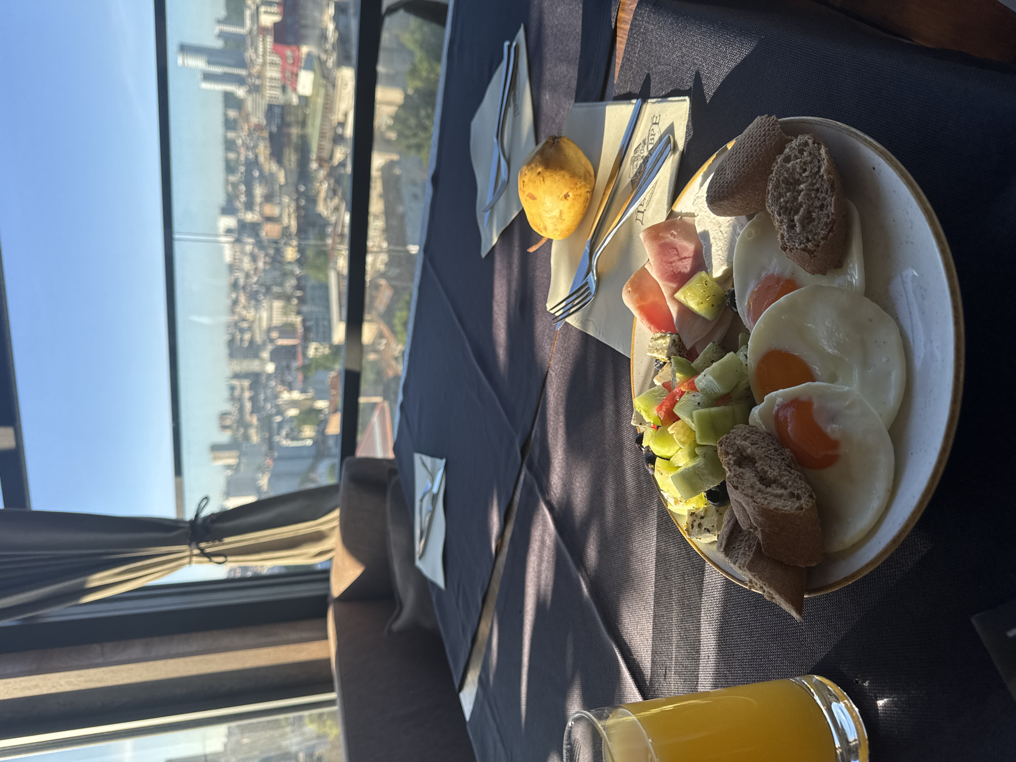 The scene is an indoor restaurant setting, likely in Belgrade, Serbia, given the skyline view. The focus is a breakfast or brunch spread on a table draped with a dark grey tablecloth. The meal includes a plate with three fried eggs, slices of dark bread, cured meats (prosciutto or ham), and a fruit salad containing diced kiwi, melon, and tomato. A whole pear rests beside the plate on a napkin, along with silverware. A glass of orange juice sits at the bottom of the frame. The background is a cityscape viewed through a large window, showing a typical urban landscape with buildings of various heights. A curtain and part of a chair are visible on the left side of the frame. Sunlight casts shadows across the table, suggesting a bright, sunny day. No people are visible in the frame.