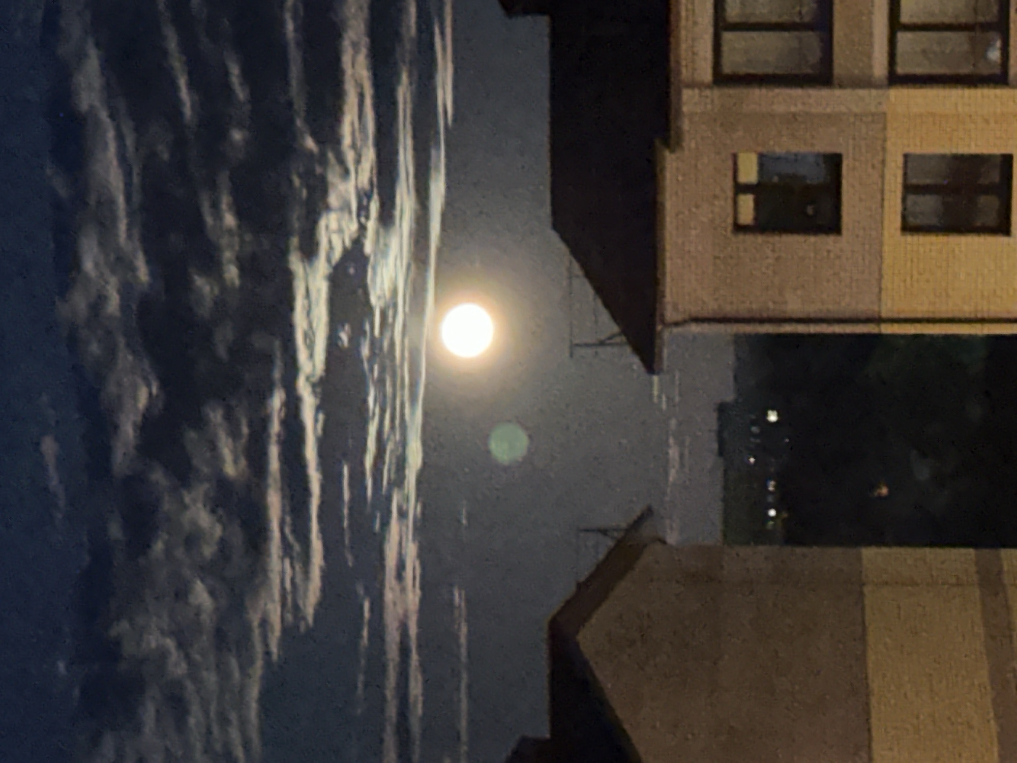 The image captures a night scene with the moon prominently visible in the sky. Wispy clouds are present on the left side of the frame. Buildings are positioned on the right, with lit windows indicating activity within. The lower portion of the image includes elements of the urban environment. The location details (city of Vladimir, Russia) are not verifiable within the frame.