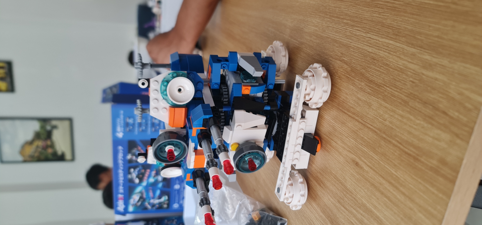 The image shows a Lego model of a vehicle, predominantly blue, white, and orange, on a light brown wooden surface. The model has wheels and what appear to be cannons or similar weapons on the front. A partial view of a person's arm is visible at the top of the image. In the background, there is a box with a blue and white design and pictures on it. Another person is partially visible in the background, to the left. The location appears to be indoors, possibly a house or apartment. There is no information in the image to determine that the location is in Đà Nẵng, Vietnam.
