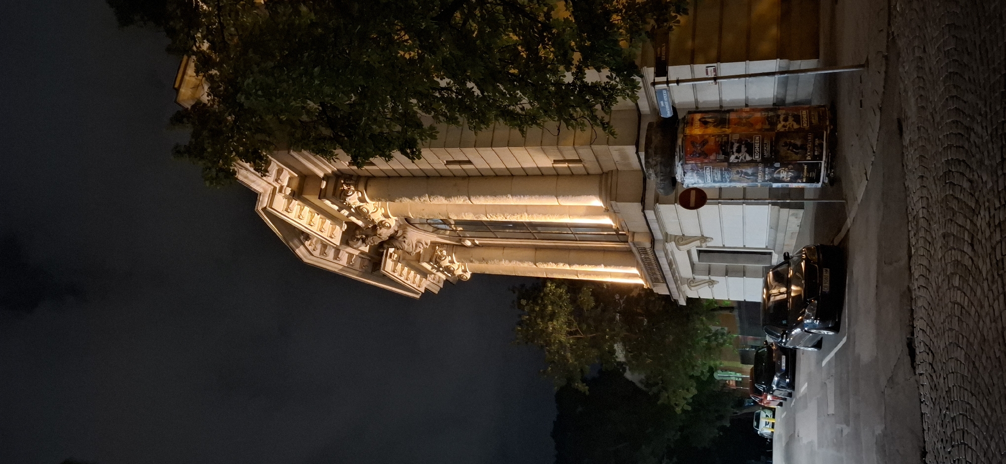 The image depicts a street scene at night in Sofia, Bulgaria, focusing on the facade of an ornate building with classical architectural elements. The building, illuminated by warm artificial lights, features fluted columns, Corinthian capitals, and decorative moldings. Tall, arched windows are visible, reflecting the exterior lighting. In the foreground, a cobblestone street runs alongside the building, with several parked cars. One car, a dark sedan, is prominently positioned, displaying a license plate that appears to read "CB 9080 HM". Next to the building, a cylindrical structure is covered with posters, some of which are partially legible, mentioning "KOSHEEN" and "BODYGUARD". A red circular "no entry" sign is also visible on this structure. Trees with dense foliage are present, partially obscuring the building and contributing to the nighttime ambiance. The sky is dark and cloudy, characteristic of an evening setting. The scene is static, with no people or active interactions apparent.