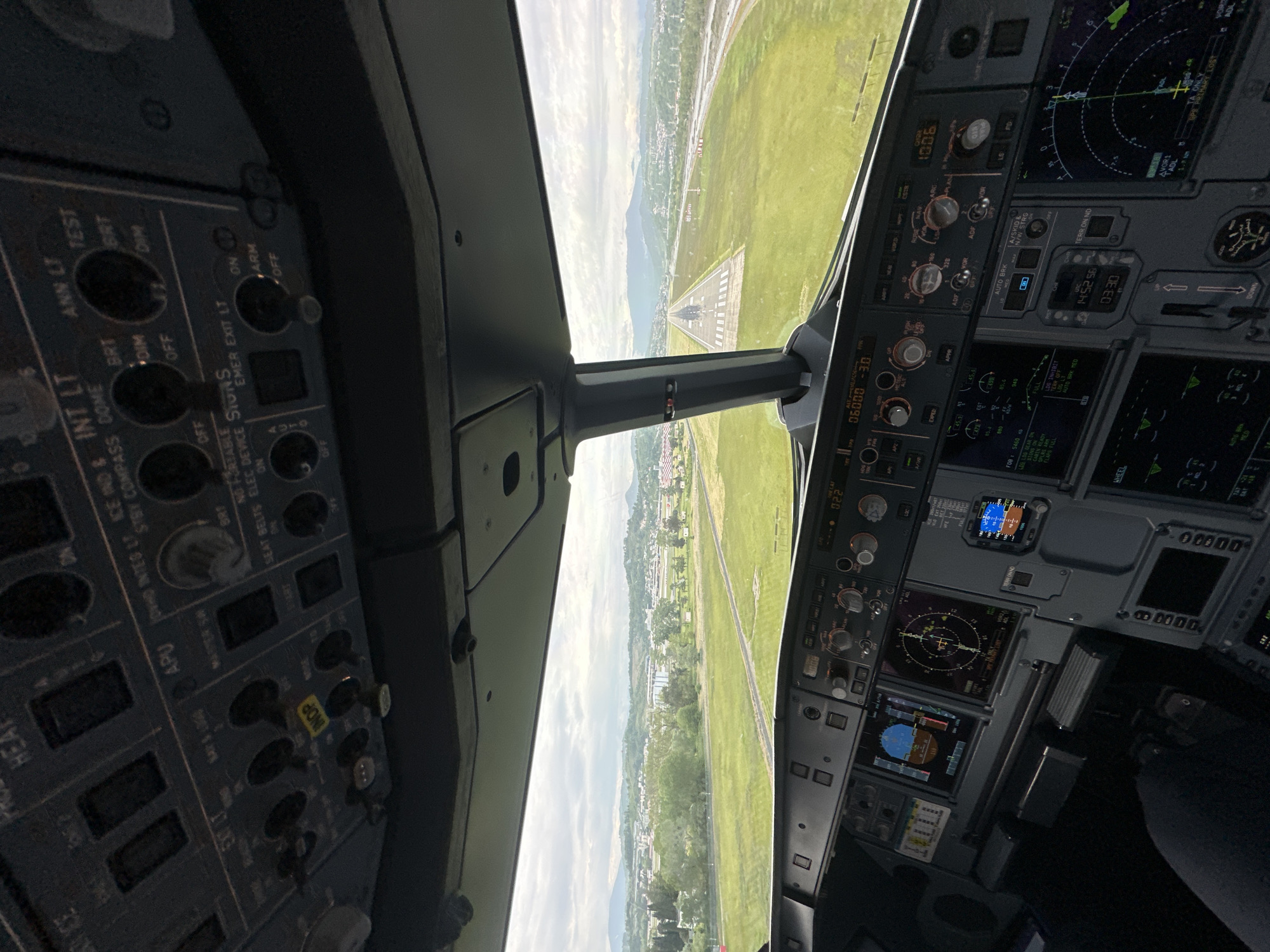 The image shows a view from the cockpit of an aircraft approaching a runway. Through the windshield, one can see a green landscape with buildings and trees, with a runway in the distance. The cockpit instrumentation is visible on both sides, including controls, displays, and gauges. Based on available details, this is not in Sochi, Russia.