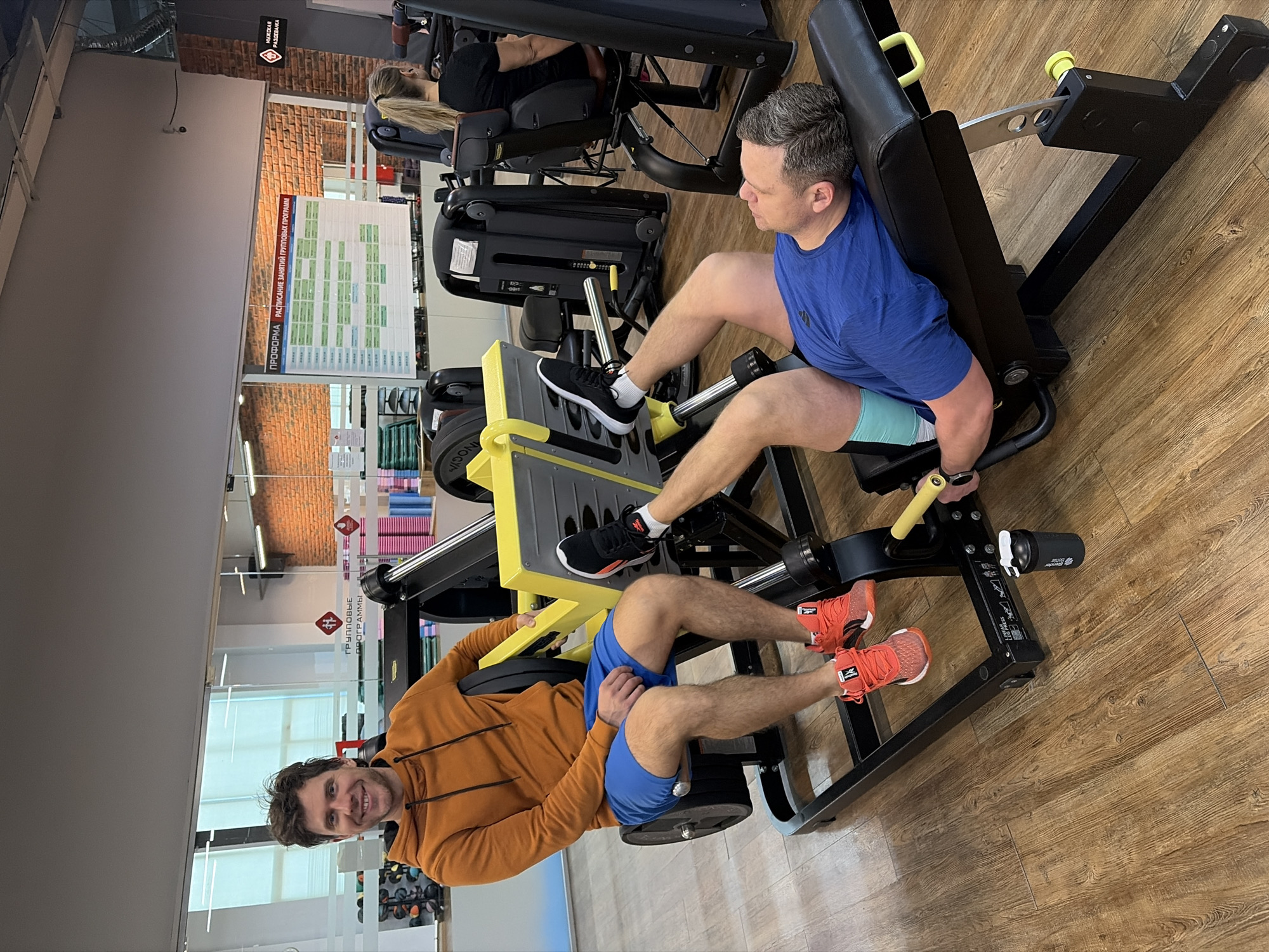 Two men are using leg press machines in a gym. The man on the left is wearing an orange hoodie and blue shorts and smiling. The man on the right is wearing a blue t-shirt and light blue shorts. They are both wearing athletic shoes. Other gym equipment and people are visible in the background. The location is Chaikovsky, Russia.