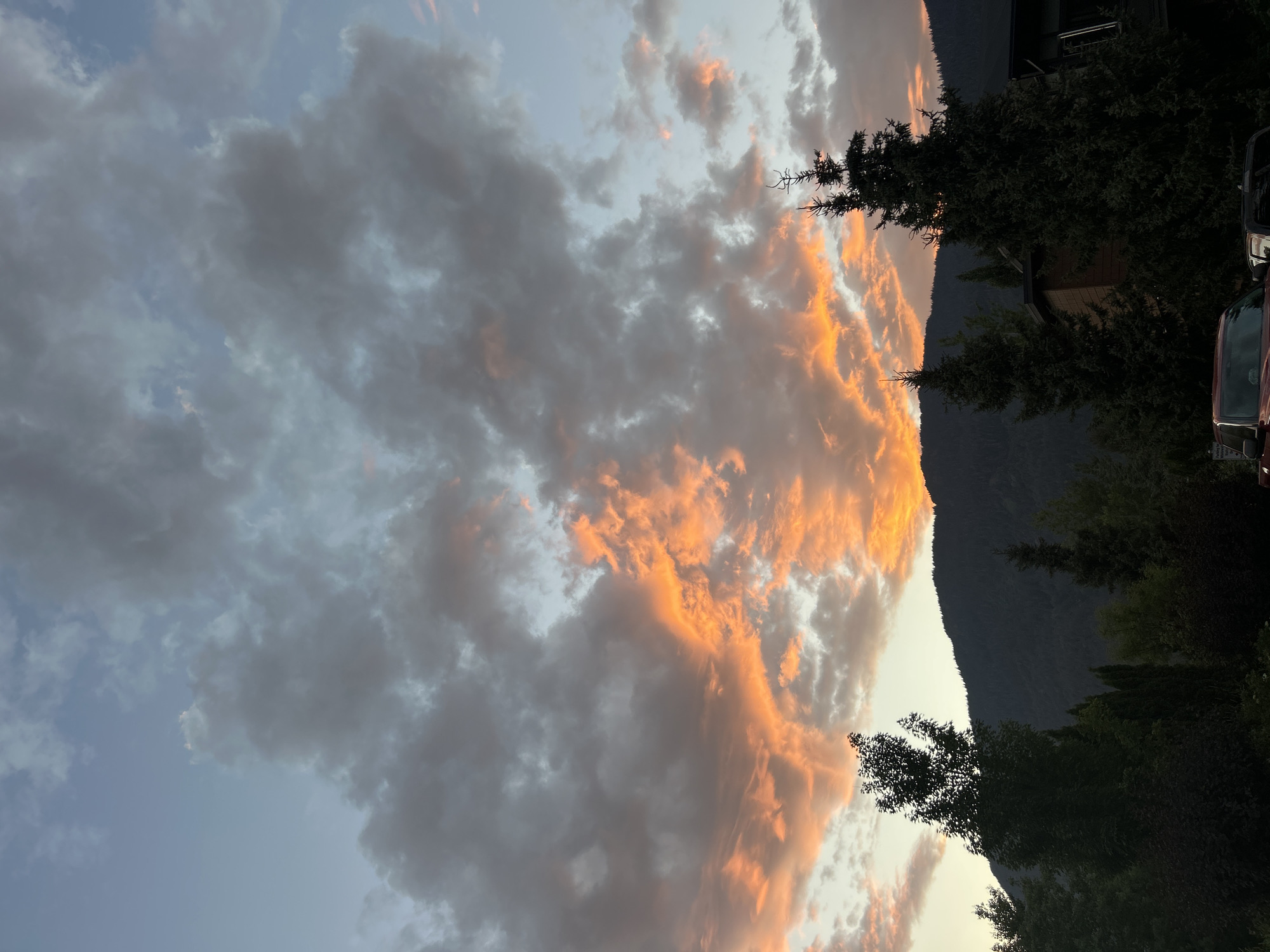 A sunset illuminates the sky with vibrant orange and pink hues, casting a warm glow on dramatic clouds. Silhouetted against the colorful sky are the dark, imposing forms of evergreen trees and the dark slopes of a mountain. In the foreground, the tops of more trees and a sliver of a building are visible. The scene is likely from Whistler, Canada, known for its stunning mountain landscapes and dramatic sunsets.