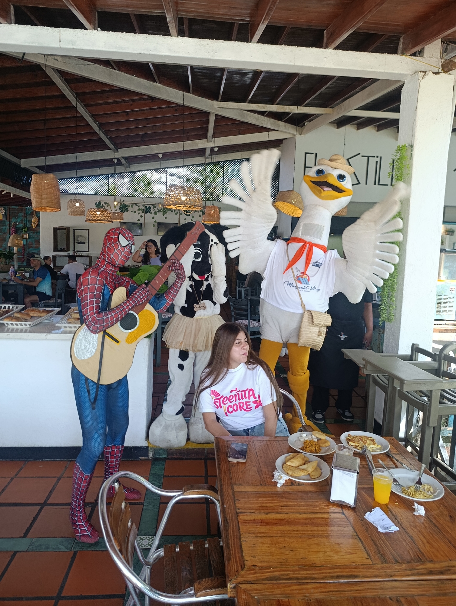 The scene is set in an outdoor restaurant in Porlamar, Venezuela, characterized by a tiled floor, wooden tables and chairs, and an exposed wooden roof structure. A young woman is seated at a table laden with several plates of food and a glass of orange juice. She is wearing a white t-shirt and jeans, and is looking to her left. Standing around her are three costumed characters: Spiderman playing a guitar, a cow, and a large bird mascot wearing a t-shirt, a hat, and carrying a small satchel. The characters are interacting with the woman, seemingly entertaining her. In the background, other people are seated at tables, and staff members can be seen.