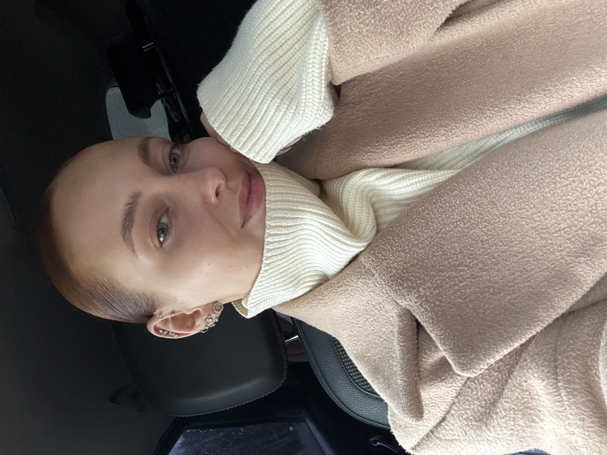 The image displays a close-up, tilted portrait of a young woman, likely taken as a selfie, within what appears to be the interior of a car. The woman has light skin, light eyes (possibly green or hazel), and her reddish-brown hair is pulled back from her face. She looks directly towards the camera with a neutral or softly contemplative expression, her lips slightly parted with a hint of gloss.
She is dressed in a cream-colored, ribbed turtleneck sweater, over which a light beige or pale pink fuzzy textured garment, possibly a coat or blanket, is draped. Her head is gently resting on her hand or arm, which is concealed by her sweater. Her visible left ear is adorned with several silver-toned piercings, including multiple small hoops and studs along the helix and lobe.
The background is dark and mostly indistinct, showing glimpses of dark, possibly leather, car seats. The lighting appears soft, suggesting an indoor setting or an overcast day. There are no other discernible objects, activities, time-of-day cues, or visible text within the frame.
