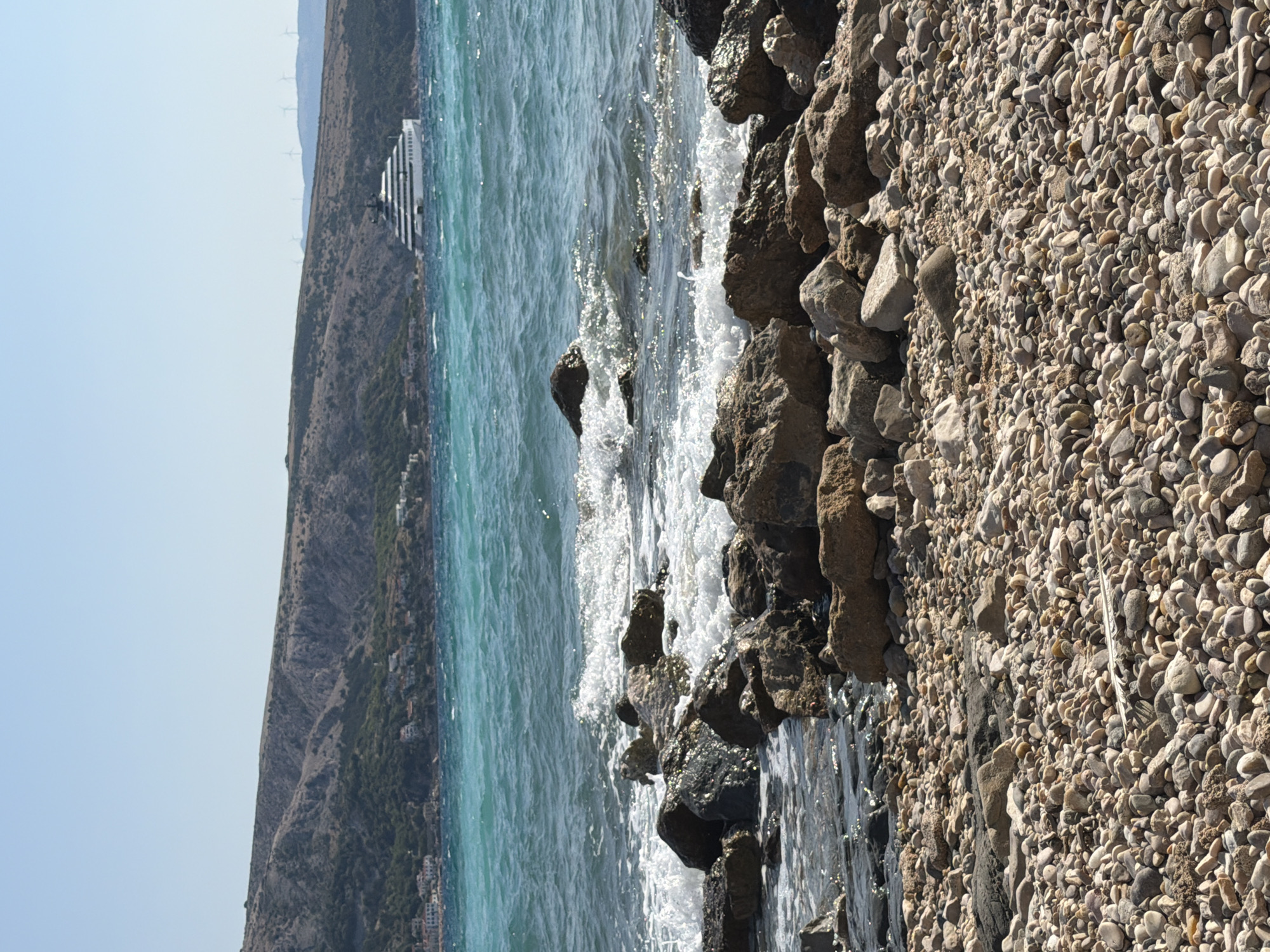 The image depicts a rocky shoreline along the coast of Općina Baška, Croatia, on a clear, sunny day. The foreground is a dense accumulation of various-sized, smooth pebbles. Immediately behind the pebbles, large, dark, wet rocks are visible, some partially submerged in the water. The sea is a vibrant turquoise, with white waves crashing against the rocks, creating sea spray and foam. In the middle ground, a large, modern white yacht with a tiered design is anchored in the water. Beyond the yacht, a sloping, arid hillside is visible, dotted with sparse green vegetation and some scattered buildings. The top of the hill features a ridge with several wind turbines. The sky is a clear, pale blue, with no clouds present.