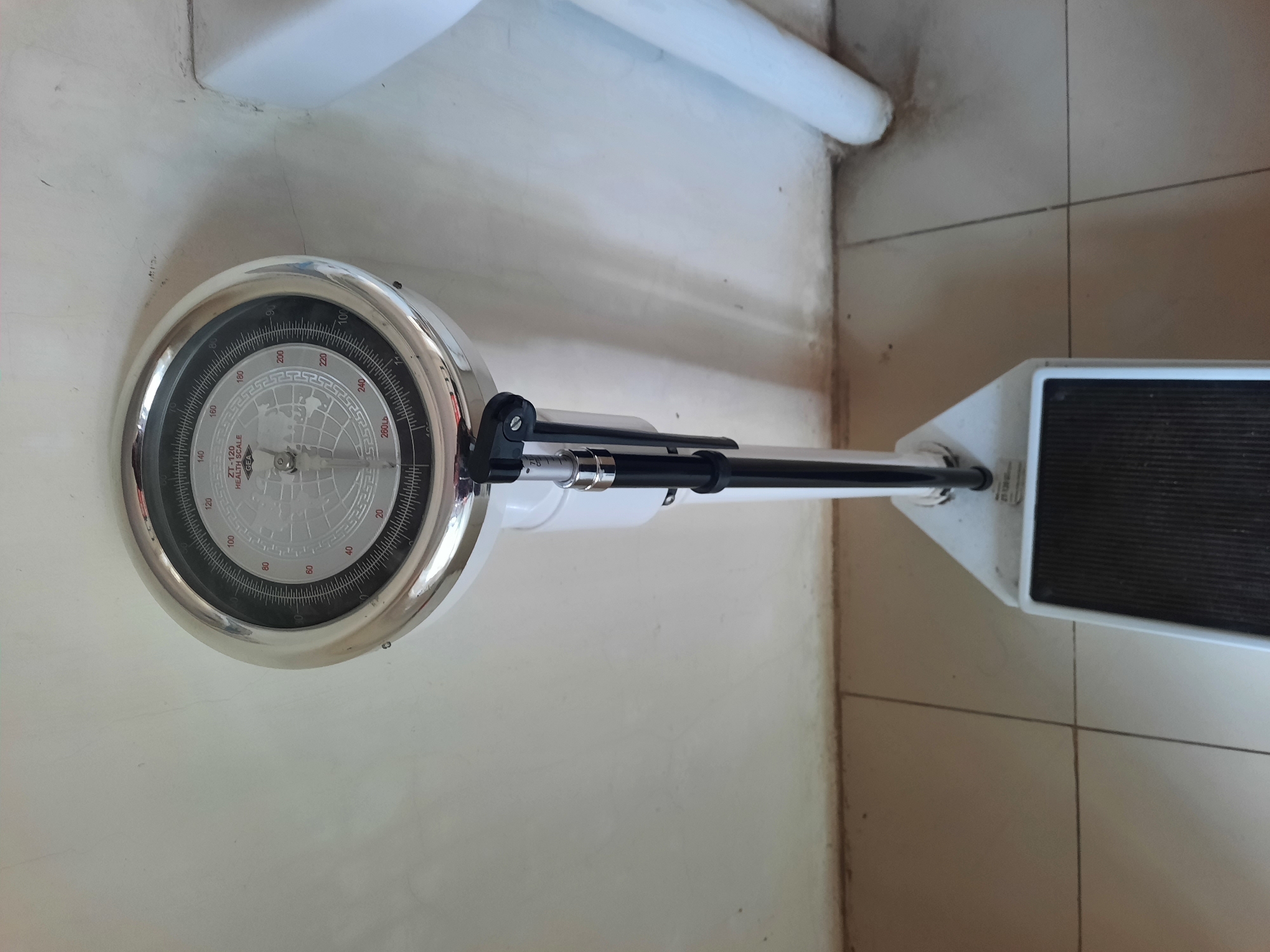 An analog health scale with a chrome-plated dial is pictured in a room with tiled flooring and a light-colored wall. The scale's dial displays weight measurements in pounds, ranging from 0 to 260 lbs, with increments of 2 lbs. The needle on the dial is positioned at approximately 120 lbs. A height measuring attachment, marked in centimeters, is extended from the scale's pole. The base of the scale is white and features a black, textured platform for weighing. The scene appears to be indoors, possibly in a clinic or personal health facility, located in the city of Lhokseumawe, Indonesia.