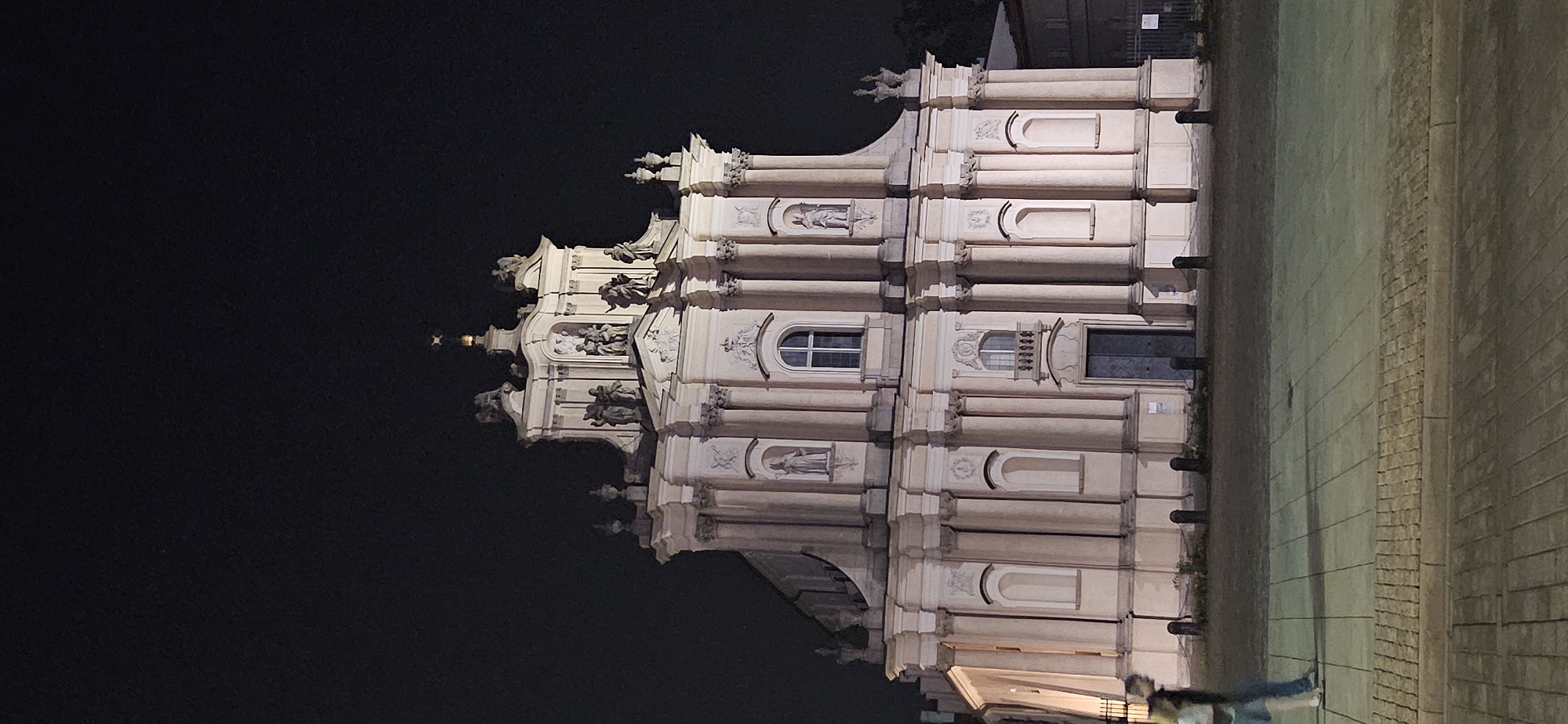 This is a nighttime photograph of the Church of the Holy Spirit in Warsaw, Poland. The Baroque-style facade of the church, illuminated by artificial lights, is the central focus. The church features classical architectural elements such as columns, pediments, and statues. The sky is dark and appears clear, with no visible celestial bodies.
In the foreground, two individuals are walking from left to right across a paved square. The person on the left is wearing a light-colored top and dark pants, while the person on the right is wearing dark clothing. They are casting shadows on the ground. The square is paved with rectangular stones, with a border of smaller cobblestones. Several dark bollards are positioned in front of the church, with a low chain connecting some of them. The overall scene depicts a quiet urban environment at night, with the grand architecture of the church dominating the view.