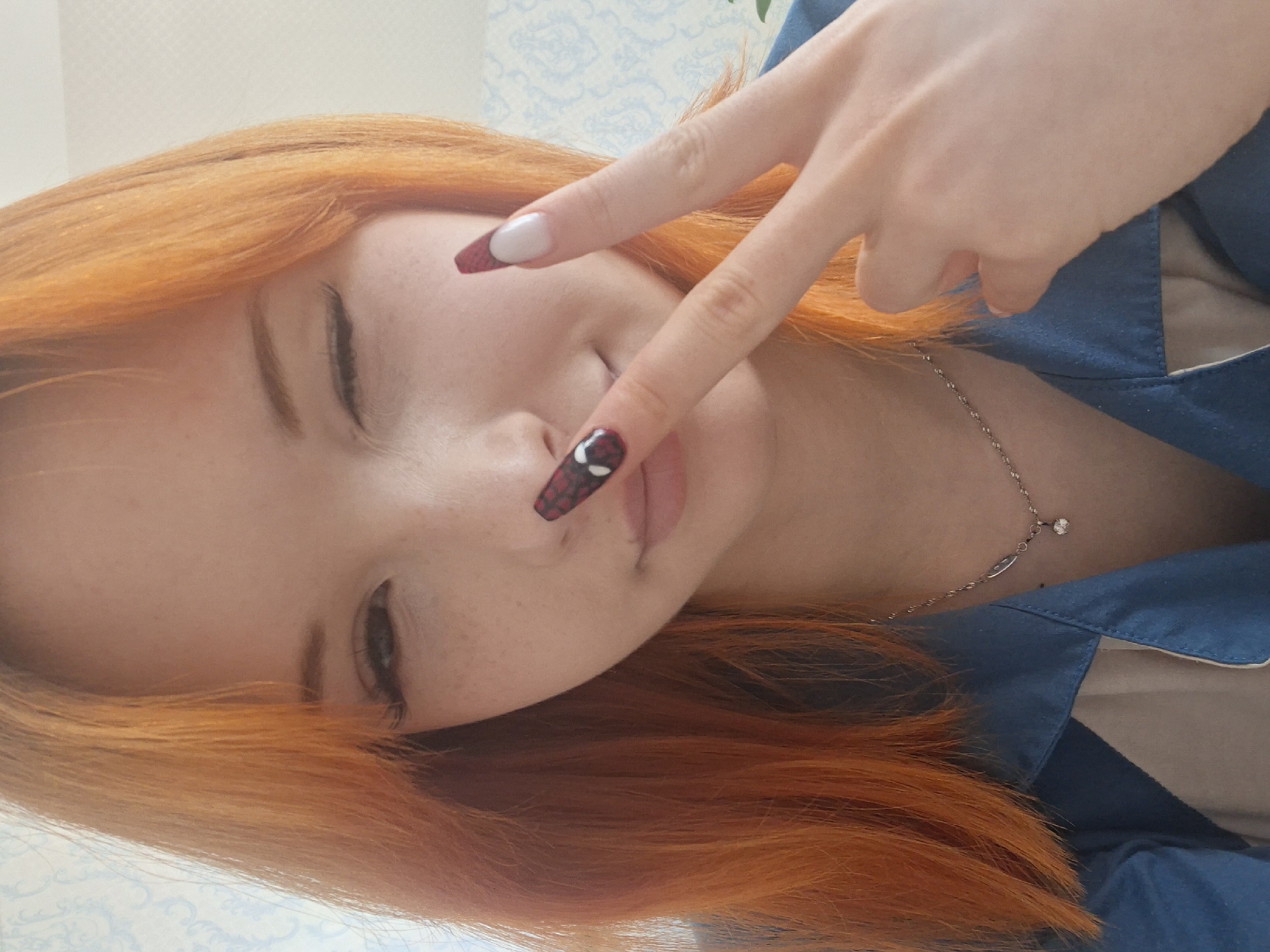 A young woman with bright orange hair poses indoors, likely in a residential setting. She is tilted to her left and looking downward. Her right hand is raised in a peace sign, with the index finger extended over her nose. Her nails are painted with a unique design: one is white with a red tip, while the other has a red and black pattern, possibly resembling a spider web or similar design. She wears a blue collared shirt and a delicate silver necklace with a small pendant. Light and wallpaper patterns are visible in the background. There is no confirmed location in Chaikovsky, Russia.