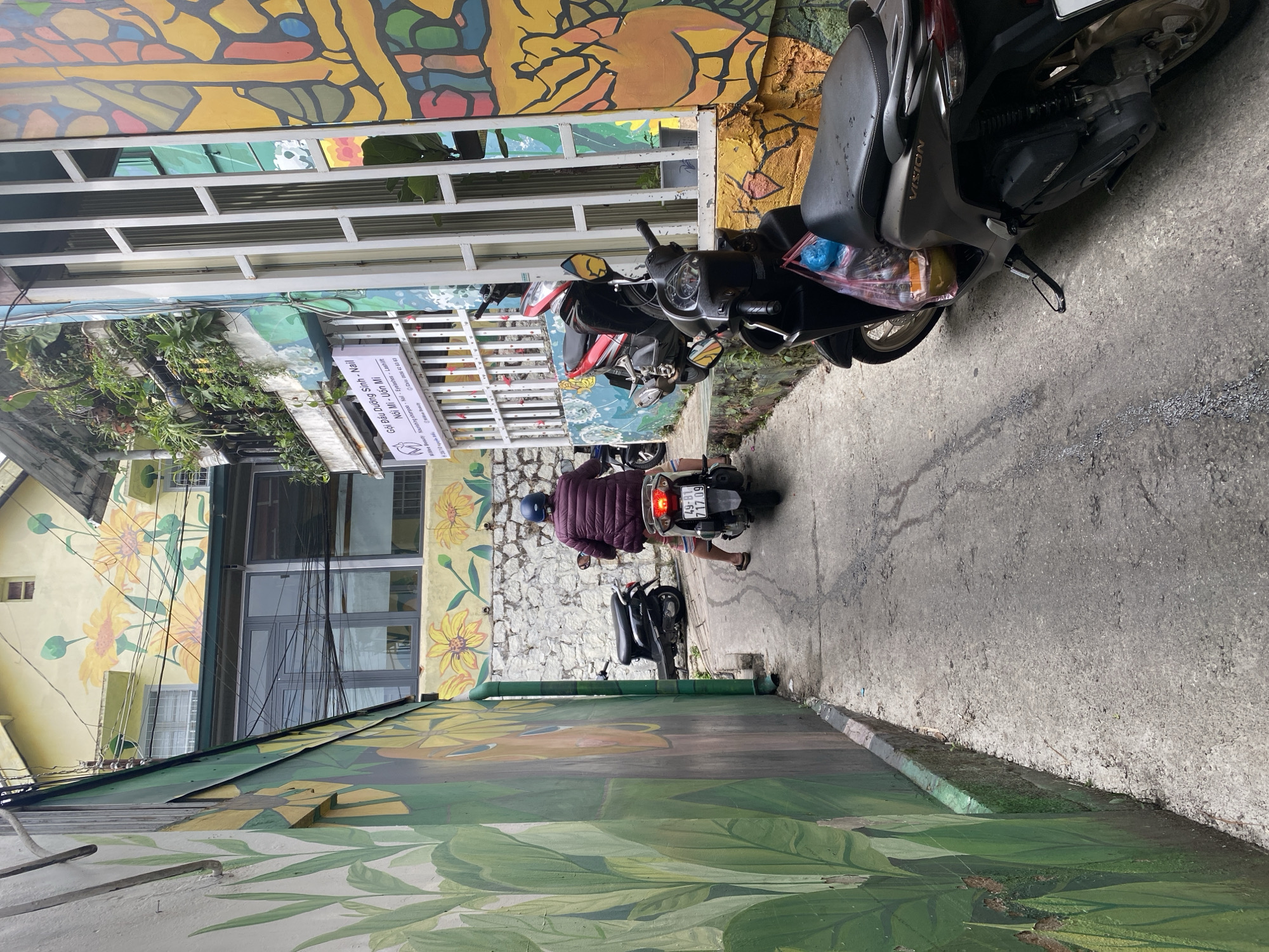 The image depicts a narrow alley in Da Lat, Vietnam. A person on a scooter, wearing a helmet and a maroon jacket, is driving away from the camera. Two motorbikes are parked on the right side of the alley. A sign in Vietnamese and English advertising a beauty service, "Gội Đầu Dưỡng Sinh - Nail" (Hair Spa - Nail), is visible. The alley walls are painted with colorful murals featuring sunflowers and abstract designs. The pavement is concrete. Overhead, there is a partially visible corrugated metal roof structure and some vegetation.