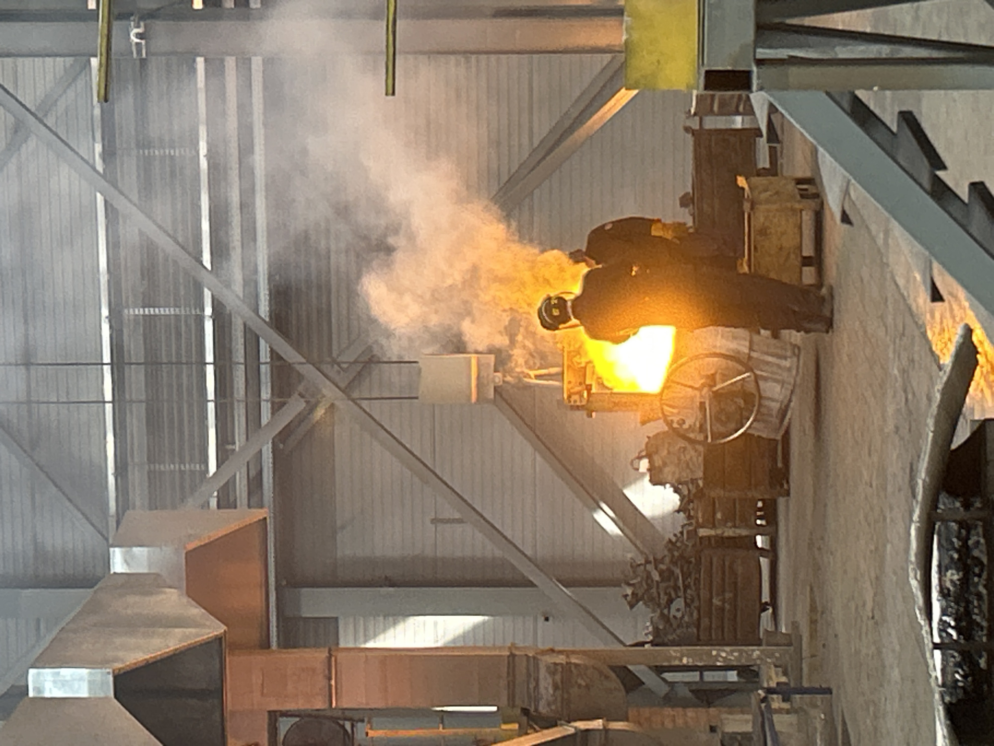 The image depicts two workers at a steel mill in Noviy, Russia. The workers are standing near a furnace, pouring molten metal into a container. Smoke is rising from the metal. The building has metal framework and is illuminated by the glow of the furnace.
