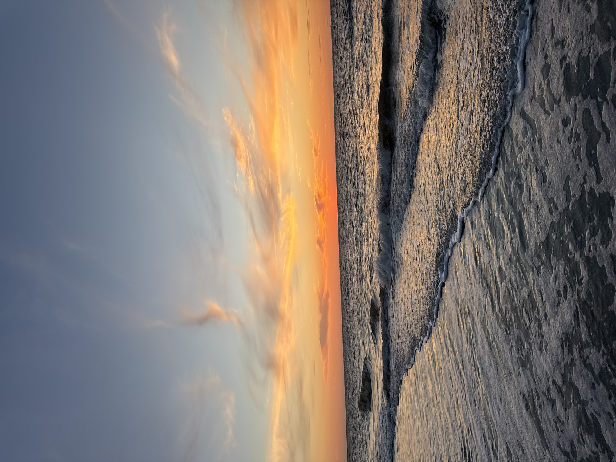The image depicts a serene beach scene at sunset on the island of Langeoog, Germany. The sky is a gradient of soft blues and vibrant oranges, with wispy clouds catching the last rays of the sun. Gentle waves are rolling onto the sandy shore, creating a foamy, white edge as they recede. The water is dark and reflects the warm colors of the sky. There are no people or artificial objects visible in the frame, emphasizing the natural beauty of the coastal environment. The overall impression is one of peaceful tranquility.