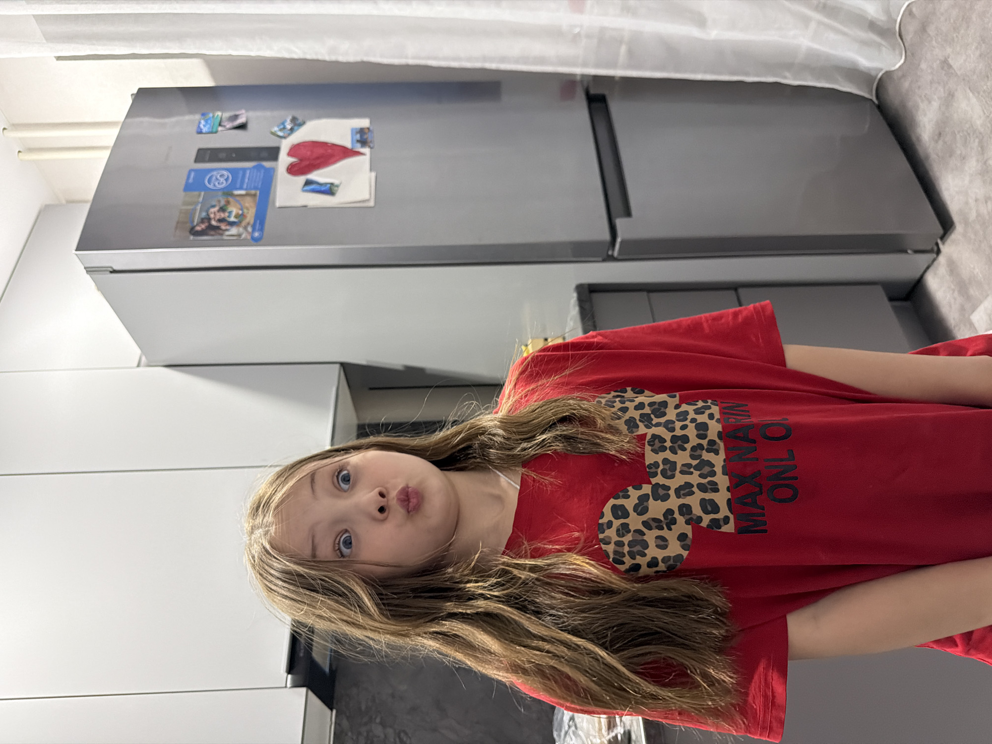 A girl with long brown hair and blue eyes stands in front of a silver refrigerator in a kitchen. The girl wears a red t-shirt with a leopard print Mickey Mouse head and the text "MAX NA RIN ONL O". The refrigerator has various magnets and papers attached to it. The kitchen has white cabinets and dark countertops. The image was likely taken indoors. There is no direct evidence the photo was taken in Chaikovsky, Russia.