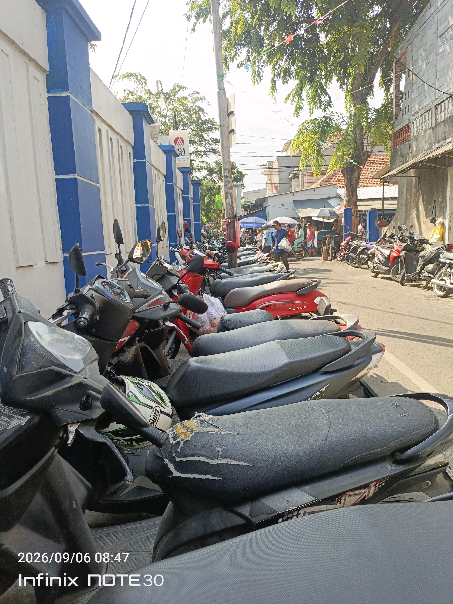 In Central Jakarta, Indonesia, a row of parked motorcycles lines a street adjacent to a white and blue building. The motorcycles are mostly scooters, with several red and black models visible. Some have helmets and bags on them. In the background, people are seen walking and shopping at stalls set up along the street. Trees with green leaves overhang the scene, and electrical wires are strung across the sky. The image is timestamped 2026/09/06 08:47 and indicates the device used is an Infinix NOTE 30.