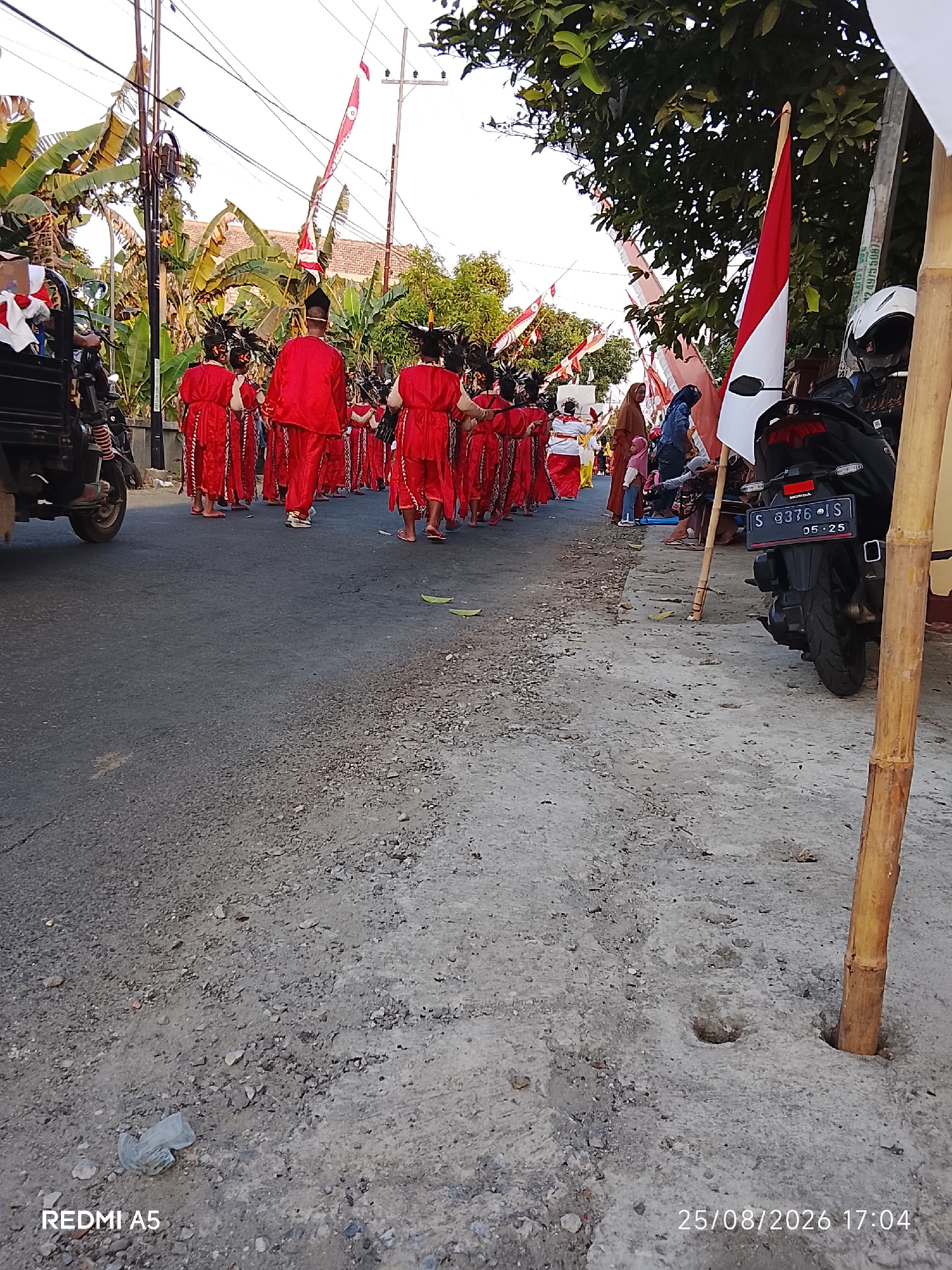 In Semanding, Indonesia, a procession is underway on a paved road. A group of people in matching red outfits with white trim, some wearing traditional headdresses, are walking in formation. Their attire suggests a cultural performance or parade. Among the onlookers are individuals in casual clothing, and a child is visible. A motorcycle with a license plate reading "S 8376 IS" is parked on the right side of the road. In the background, trees and buildings can be seen. A red and white flag, likely the Indonesian flag, is being carried. The ground is a mix of asphalt and gravel, with some scattered debris. The timestamp "25/08/2026 17:04" is visible in the lower right corner, suggesting the image was taken on August 25, 2026, at 5:04 PM. The "REDMI A5" watermark indicates the device used to capture the image.