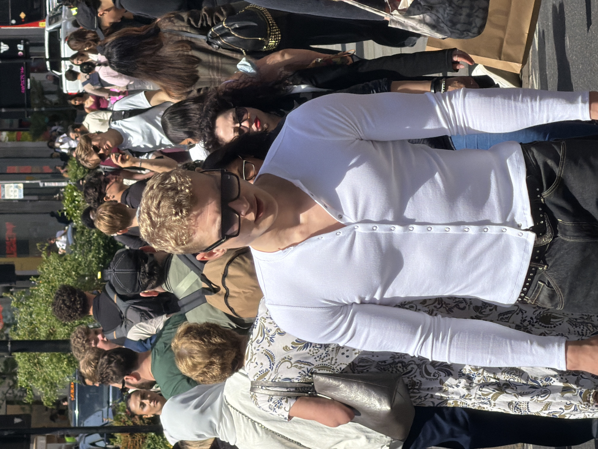 The image depicts a street scene in London, United Kingdom. A dense crowd of people occupies the frame.
Foreground focus features a man in a white, long-sleeved, button-up shirt and black jeans with decorative embellishments. He is wearing sunglasses and has blond hair. Adjacent to him is a woman in a patterned white top with a silver purse. Behind them, a woman with dark hair wears red lipstick and sunglasses, wearing a black leather jacket.
The background features a diverse group of people, some wearing sunglasses and varying styles of clothing. A vehicle and building signage are visible in the blurred background. The lighting suggests a sunny day.