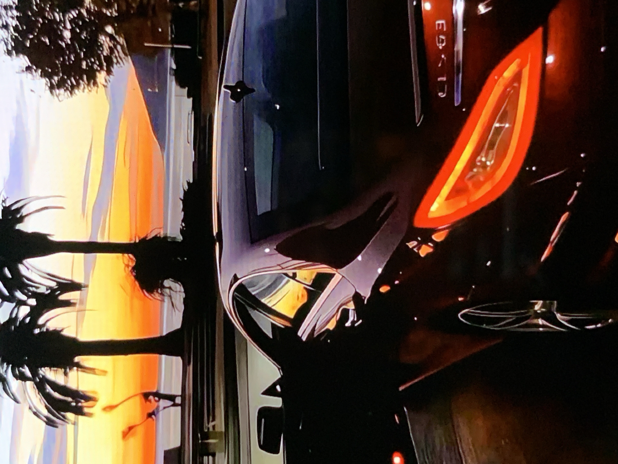 A dark-colored sedan is parked in front of a sunset scene. The car's rear taillight is illuminated with a vibrant red and orange glow. The sunset is painted with shades of orange, yellow, and purple, with silhouetted palm trees framing the scene. A body of water is visible in the distance, with a mountain range in the background. A person is standing on a pier in the distance, silhouetted against the colorful sky. The car appears to be parked on a paved surface, possibly a street or parking lot, with a line of dark objects that could be benches or railings in the foreground. The car's emblem, "CLS63," is visible on the trunk.