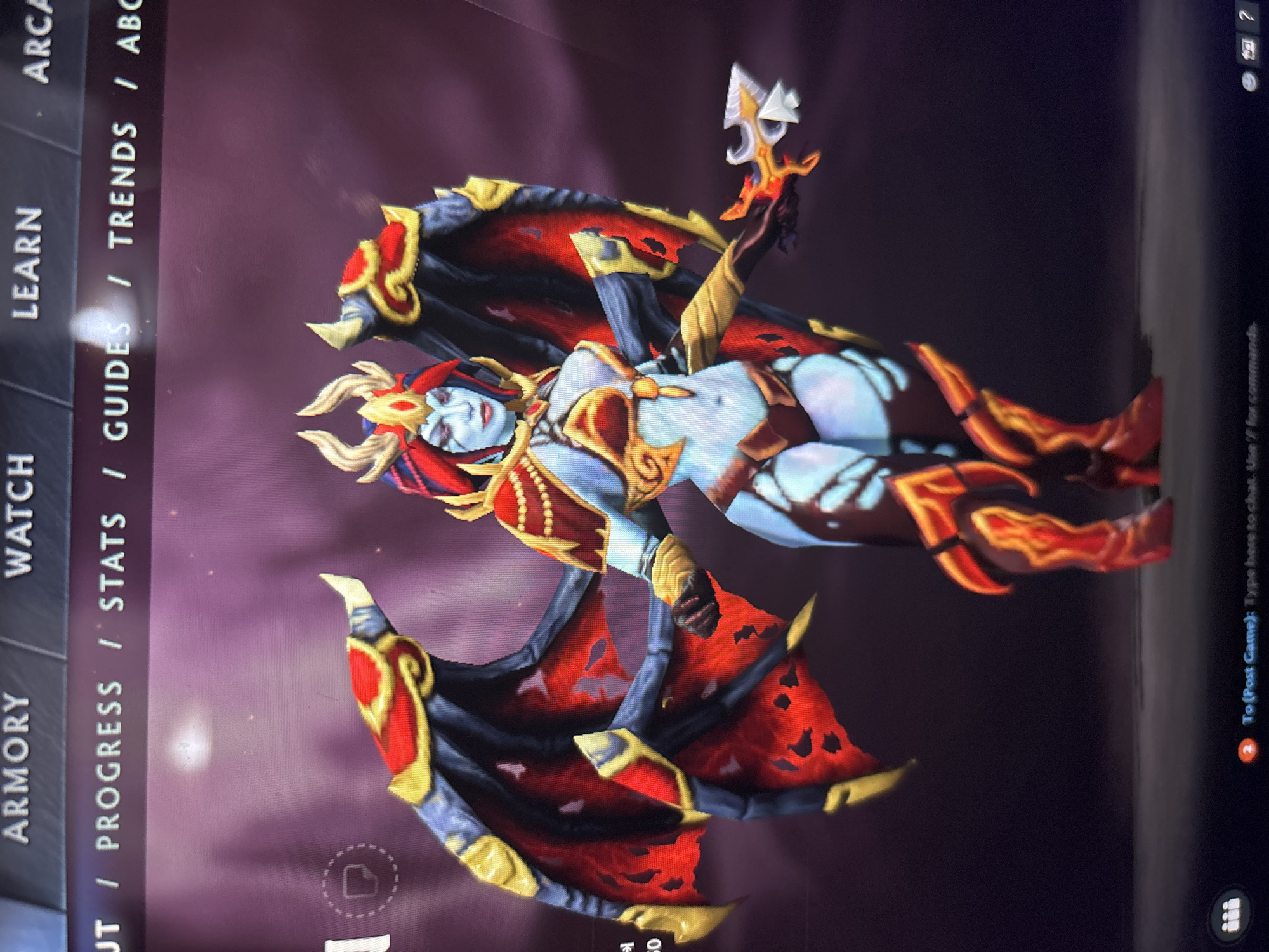 A vertical image captures a computer screen displaying a video game character and interface elements. The central subject is a stylized female character, likely from a fantasy game, with light blue skin, red and gold attire, horns, and large, tattered red and black wings. She holds an ornate, flaming dagger in her raised right hand. Her lower body also features fiery visual effects. The character appears to be a demon or succubus, posed dynamically against a dark, blurred background that suggests an in-game environment or character showcase screen.
The screen's left side features menu options oriented vertically due to the image's rotation. Visible text includes "ARMORY," "WATCH," "LEARN," and sub-categories such as "/ PROGRESS," "/ STATS," "/ GUIDES," and "/ TRENDS." A partially visible "ARCA" is at the top right. In the bottom right corner of the screen, a chat or command prompt reads "To (Post Game): Type here to chat. Use '/' for commands." The character's design, particularly her appearance and wings, is indicative of the "Queen of Pain" hero from the game Dota 2. The image appears to be a photograph of a screen in Sofia, Bulgaria.