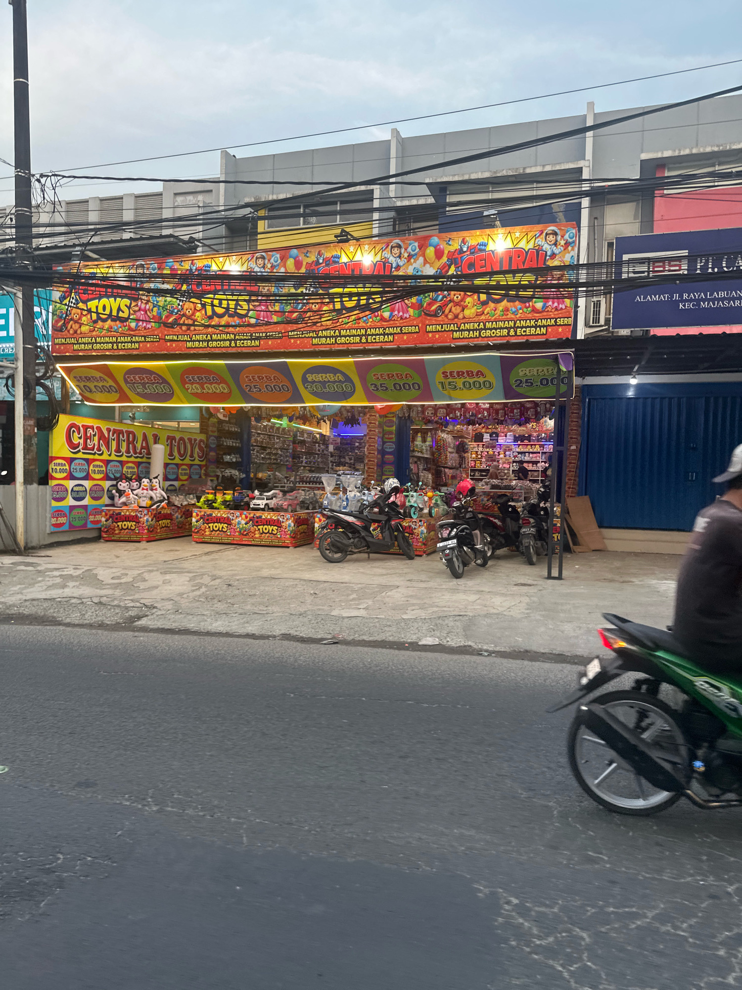 This is a street-level, medium-distance, eye-level shot of a toy store called "CENTRAL TOYS" in Saruni, Indonesia. The store is brightly decorated with banners featuring cartoon characters and text in Indonesian, advertising "ANAK-ANAK SERBA MURAH GROSIR & ECERAN" which translates to "Children's Everything Cheap Wholesale & Retail". The store appears to sell a variety of toys, with shelves packed with merchandise visible inside. In front of the store, there are several displays of toys, including plush animals and toy vehicles. Several motorcycles are parked on the sidewalk in front of the store, and a person is riding a motorcycle in the foreground, partially obscuring the view of the store. The street in front of the store is paved and appears to be a busy thoroughfare. The sky is overcast.