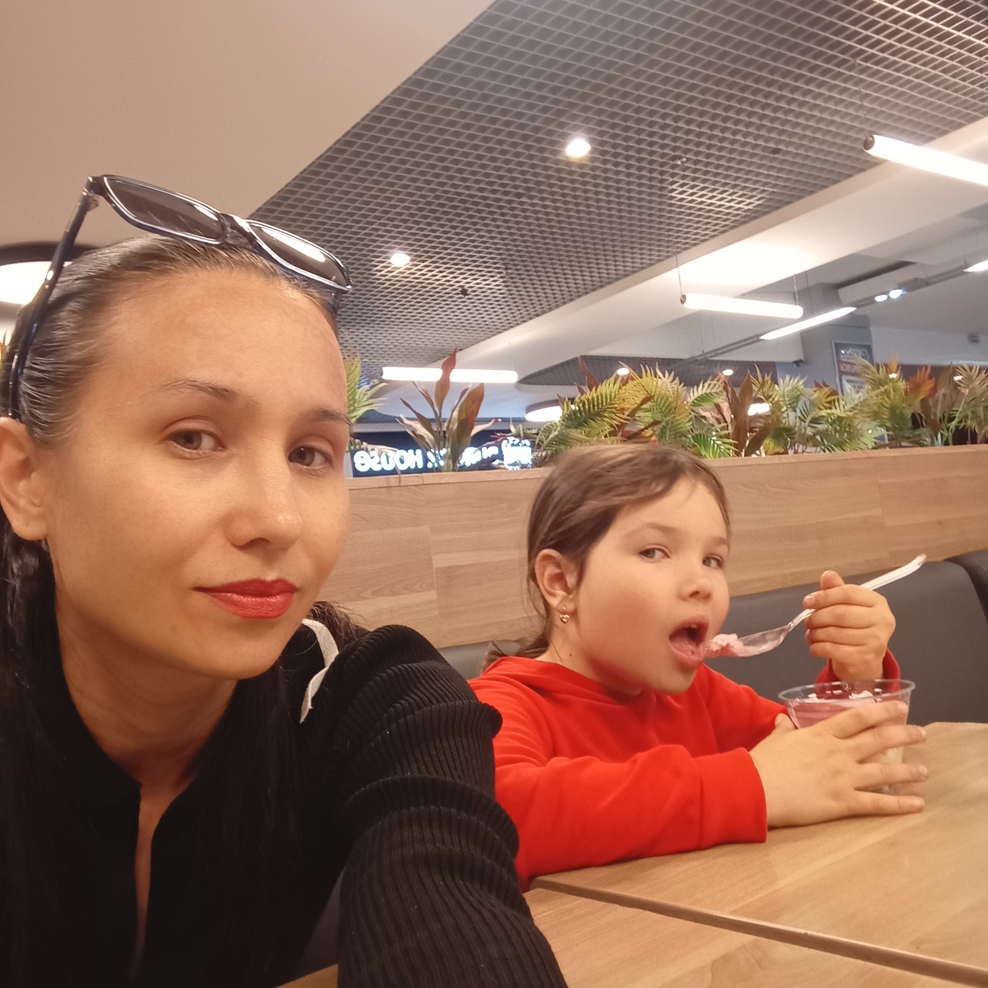 A selfie is taken by an adult woman with long dark hair, wearing sunglasses on her head and a black top with a white stripe. She is in the foreground, looking to her left. To her right, a young girl with brown hair, wearing a red hoodie, is eating ice cream from a plastic cup with a spoon. She is looking forward with her mouth open. They are sitting at a wooden table in what appears to be a food court or restaurant, indicated by the background which includes a ceiling with lights and a grid structure, potted plants, and a blurred sign that says "e200H". The overall setting suggests an indoor public eating area.
