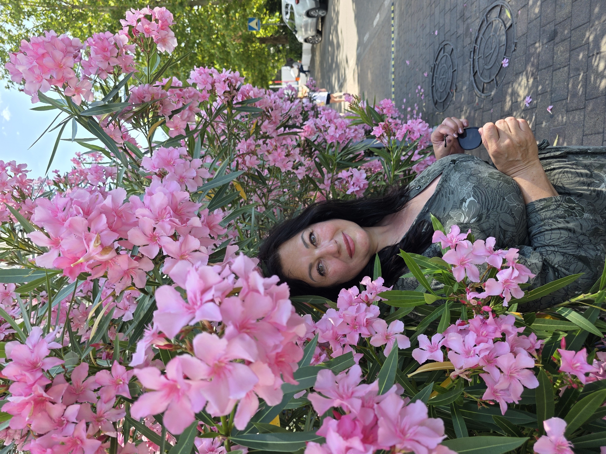 A middle-aged woman with long dark hair and light skin is standing next to a dense bush of pink oleander flowers. She is wearing a long-sleeved, dark green dress with a floral pattern and appears to be looking at the camera with a slight smile. She is holding a pair of dark sunglasses in her right hand. The background shows a paved area with manhole covers, a sidewalk, and trees. In the distance, there are cars and people, suggesting an outdoor, urban setting. The lighting indicates it is daytime, likely sunny given the shadows and bright sky. The image is taken at a slight upward angle. The scene is not specifically identifiable as Sochi, Russia, but rather a general outdoor urban location.