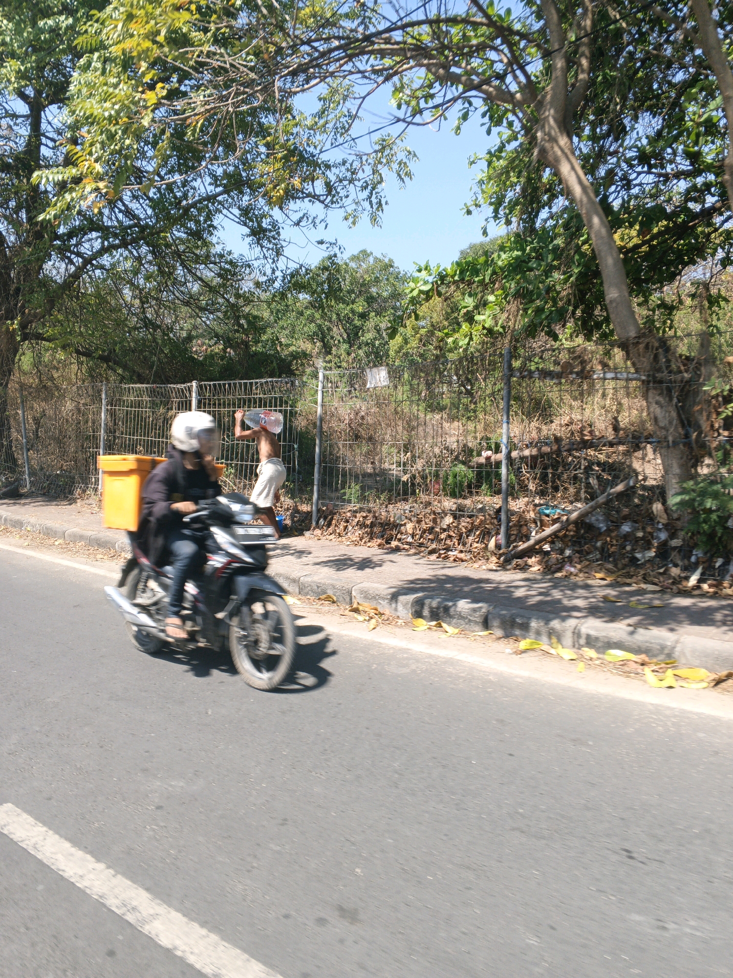 In Kuta, Indonesia, a person is riding a motorcycle with a yellow container on the back. Another person is visible behind a fence, carrying a large plastic bottle on their shoulders. The environment consists of trees, a fence, and a road with a curb. The scene appears to be outdoors on a sunny day.