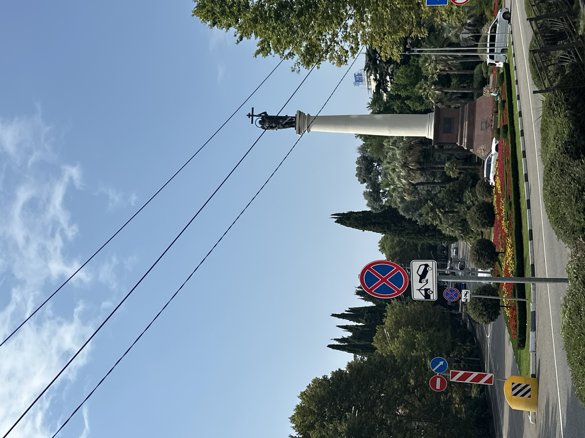 The image depicts a street scene in Sochi, Russia, dominated by a tall white column topped with a statue of a figure holding a cross. Several power lines stretch across the sky. In the foreground, there are traffic signs, including a "no stopping" sign and a sign indicating a tow-away zone. A circular flower bed with red and yellow flowers is visible to the right of the road, with a white SUV and a white minivan parked nearby. Trees, including palm trees and cypress trees, surround the area. The sky is clear and blue with a few scattered clouds.