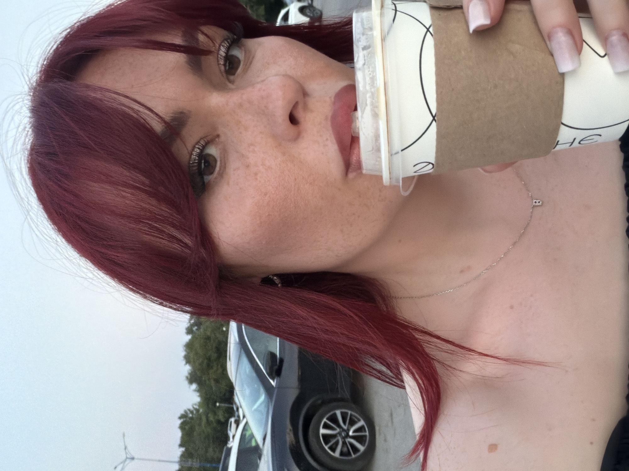 A medium close-up photograph captures a young woman with red hair and light skin, wearing a black top, as she sips from a disposable coffee cup. She has noticeable freckles across her nose and cheeks, and her makeup includes winged eyeliner and light pink lipstick. A delicate silver necklace with a small pendant is visible around her neck. Her fingernails are painted a light pink. She is holding the paper cup with a brown cardboard sleeve. In the background, several cars are parked in what appears to be a parking lot, with trees and streetlights visible further in the distance. The sky is a pale blue, suggesting it is either early morning or late afternoon. The location is Tolyatti, Russia, based on the presence of distinct markings on the coffee cup consistent with local branding.