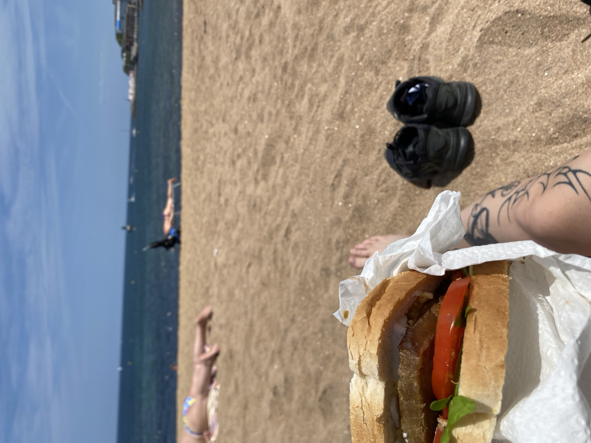 The image shows a person sitting on a sandy beach, possibly in Gijón/Xixón, Spain, as the shoreline appears to have a city visible in the distance. The sky is clear and blue, and the sea is a deep blue color. In the foreground, a partially eaten sandwich wrapped in white paper rests near the person's legs, one of which has a tattoo. Black shoes are placed nearby on the sand. Further out, other individuals are visible in the water. The composition suggests a relaxed beach setting during the daytime.