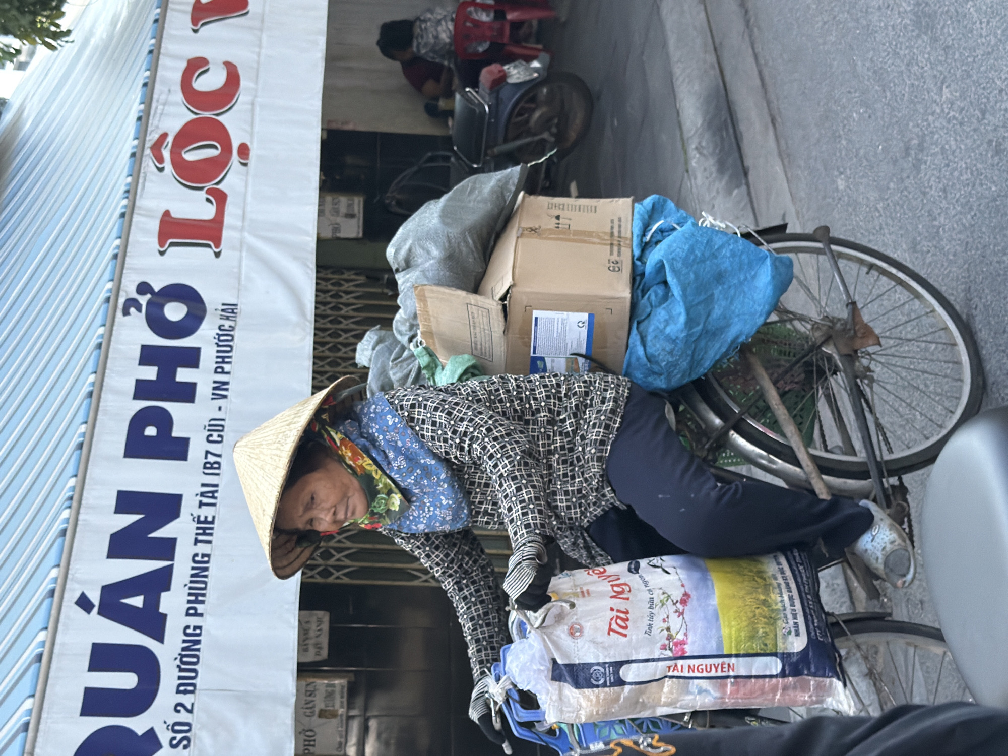 A woman wearing a conical hat and patterned shirt rides a bicycle loaded with goods in front of a building with a striped awning in Nha Trang, Vietnam. The awning has Vietnamese text in large blue and red letters. The woman has a scarf around her neck and gloves on her hands. The bicycle is carrying several bags, boxes, and a blue tarp. In the background, a motorcycle is parked, and two people are seated at a table. The scene appears to be on a street with a sidewalk.