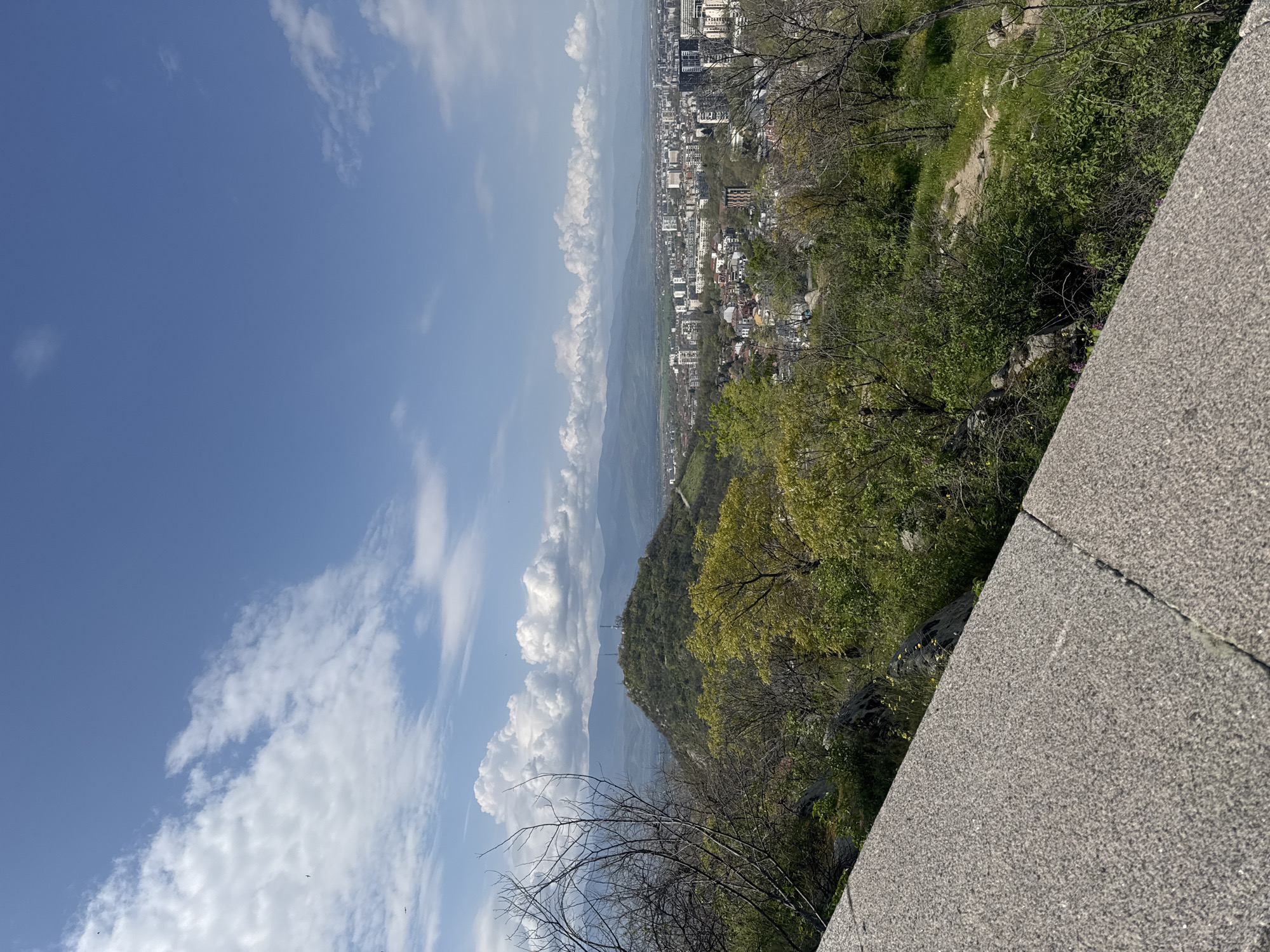The image shows a view from Nebet Tepe in Plovdiv, Bulgaria, looking out over the city. In the foreground, a concrete retaining wall is visible. A mix of trees and vegetation partially obscures a view of the city below. In the distance, more of the cityscape is visible, with buildings varying in height. The sky is blue with scattered clouds, some forming a vertical band on the left side of the image. The viewpoint is elevated, suggesting a location on a hill.