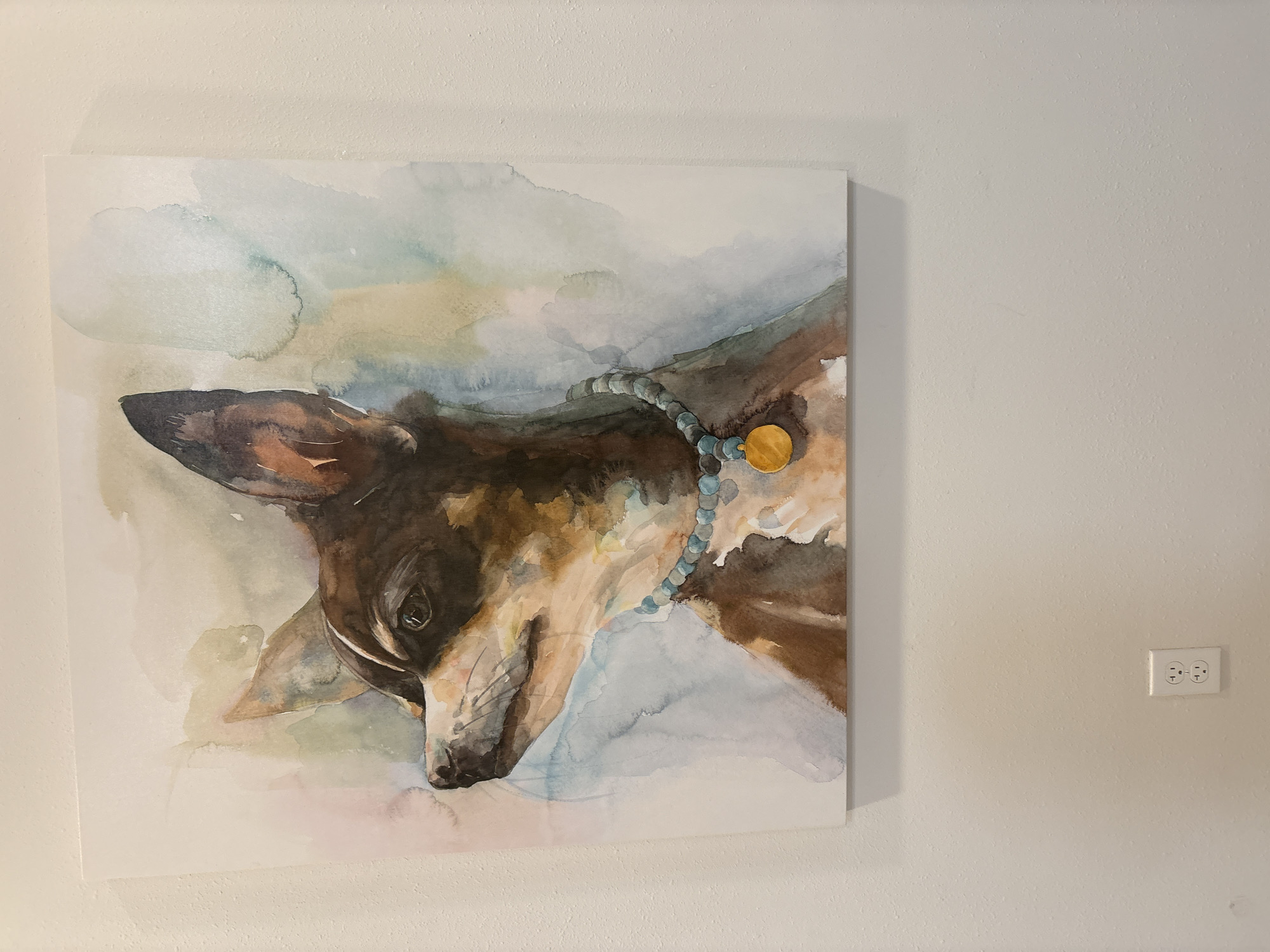 A watercolor painting of a dog is hanging on a textured, off-white wall. The painting depicts a chihuahua or similar small dog with brown, black, and white markings, looking to the left. The dog is wearing a necklace of blue beads and a gold pendant. The background of the painting is a wash of soft blues, greens, and pinks. Below the painting, centered on the wall, is a standard white electrical outlet. The overall scene appears to be an indoor residential setting in Lakewood Ranch, United States.