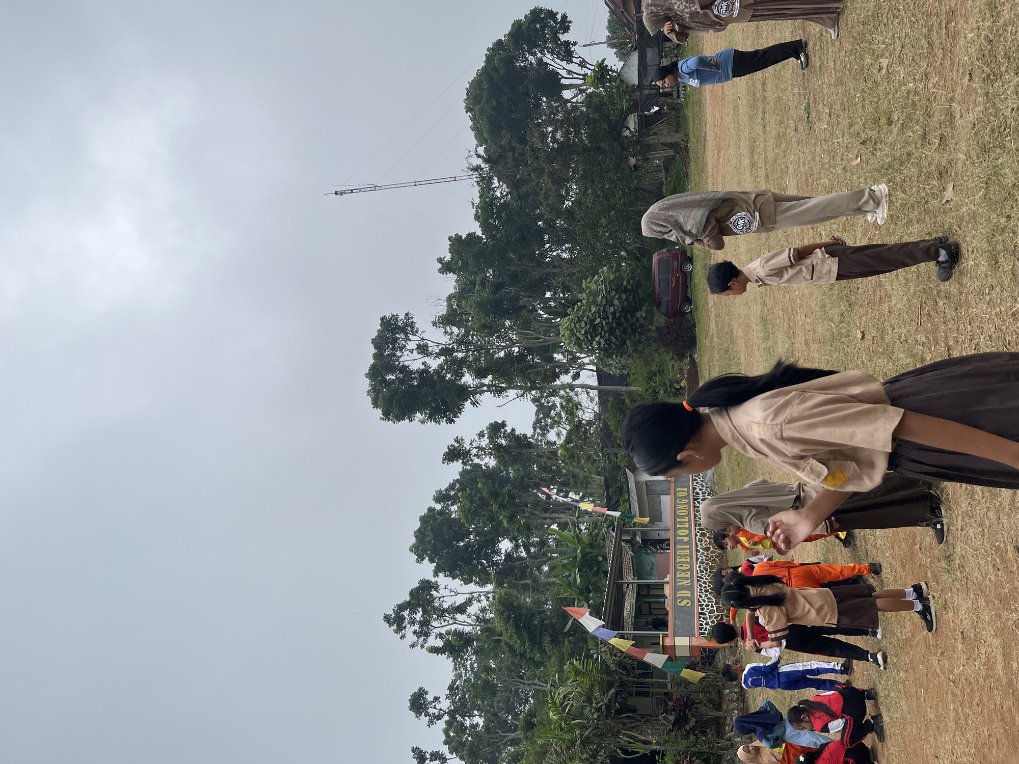 The image depicts an outdoor scene at SD Negeri Jollong 01 in Gembong, Indonesia. A group of children in school uniforms, some in brown and beige, others in blue and red, are gathered on a grassy field. Some children appear to be engaged in an activity, possibly a physical education class or a school event, with several standing in formation or interacting. A few adults are also present, observing the children. In the background, a building with "SD NEGERI JOLLONG 01" visible on its facade stands amongst lush greenery and tall trees. A red van is parked to the left of the building, and a tall communications tower is visible in the sky. The sky is overcast.
