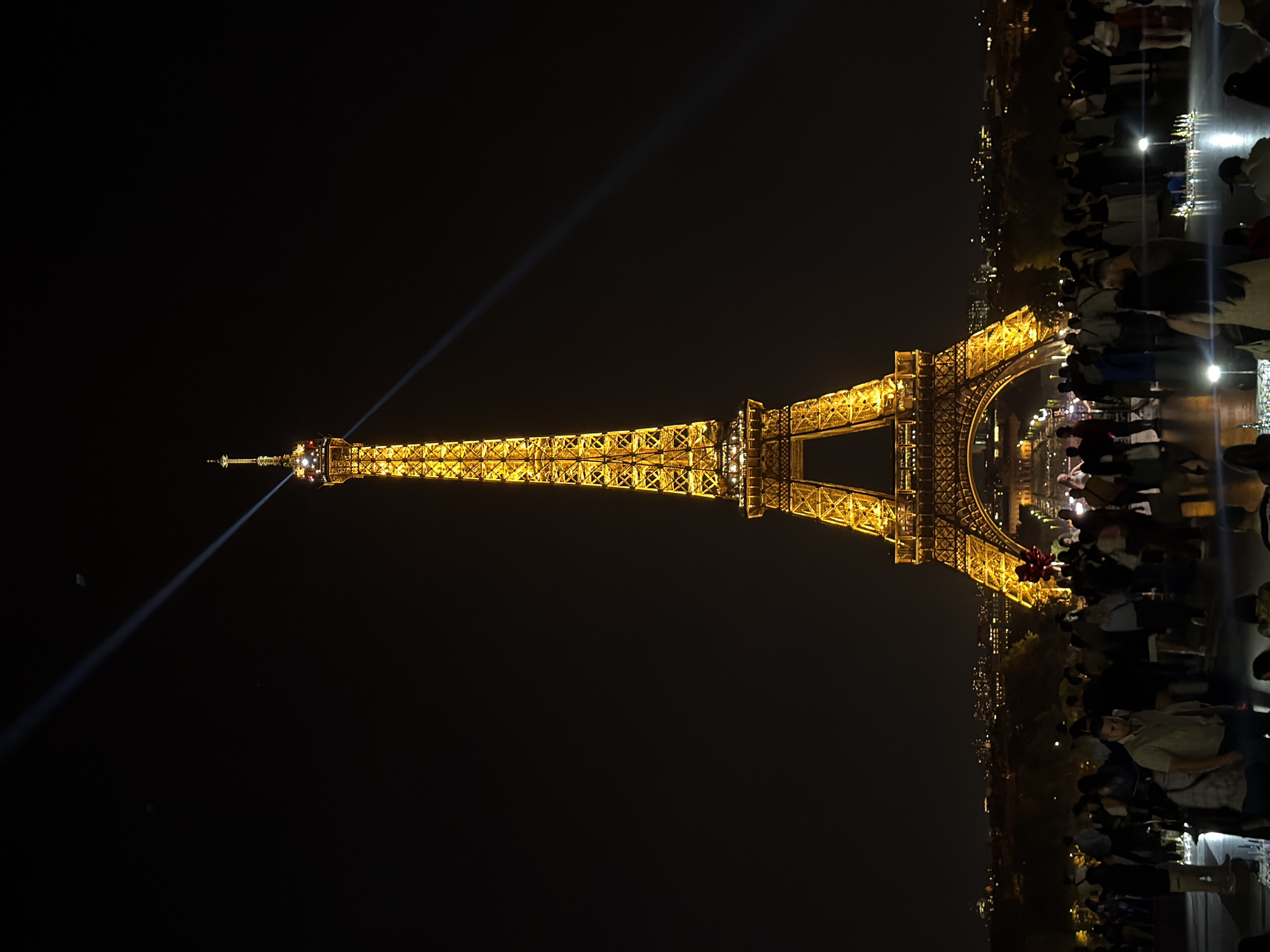 At night, the Eiffel Tower in Paris, France, stands illuminated against a dark sky, with a distinct beam of light emanating from its peak. Crowds of people are gathered in close proximity to the tower. The surrounding cityscape is visible with numerous lights.