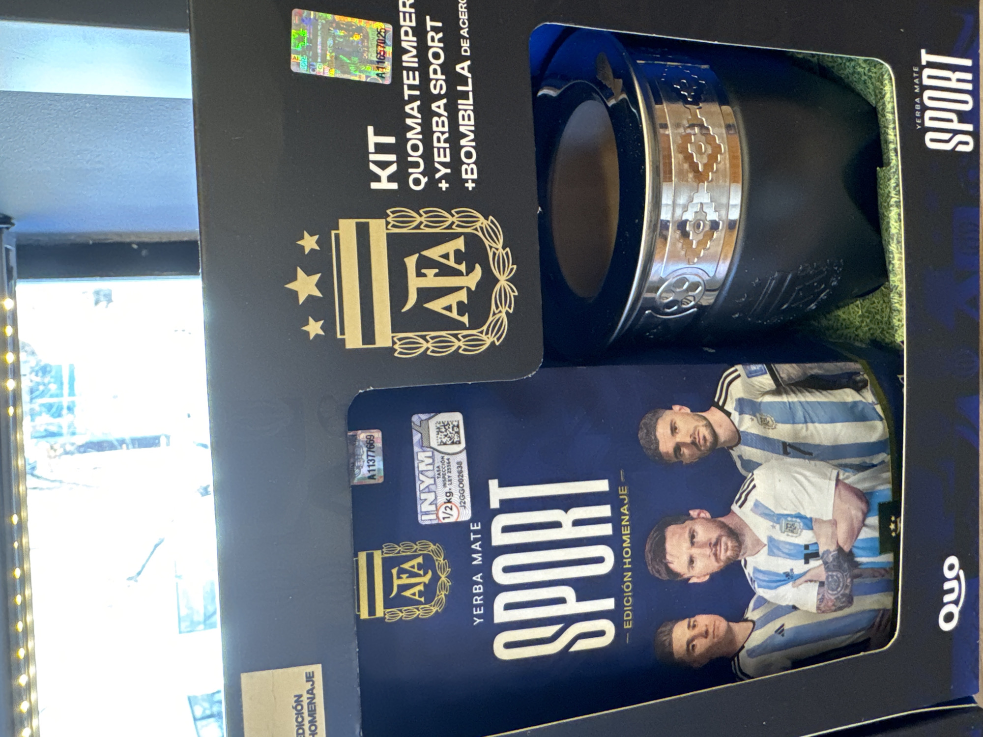 The image shows a product box, with a kit that includes a QUOMATE, YERBA SPORT and a BOMBILLA DE ACERO. There's also a bag of yerba mate, sporting three soccer players (presumably from Argentina) wearing the national team jersey. The kit appears to be an homenaje edition, and has logos and mentions of AFA (Argentine Football Association). It's in Buenos Aires, Argentina based on the AFA association.