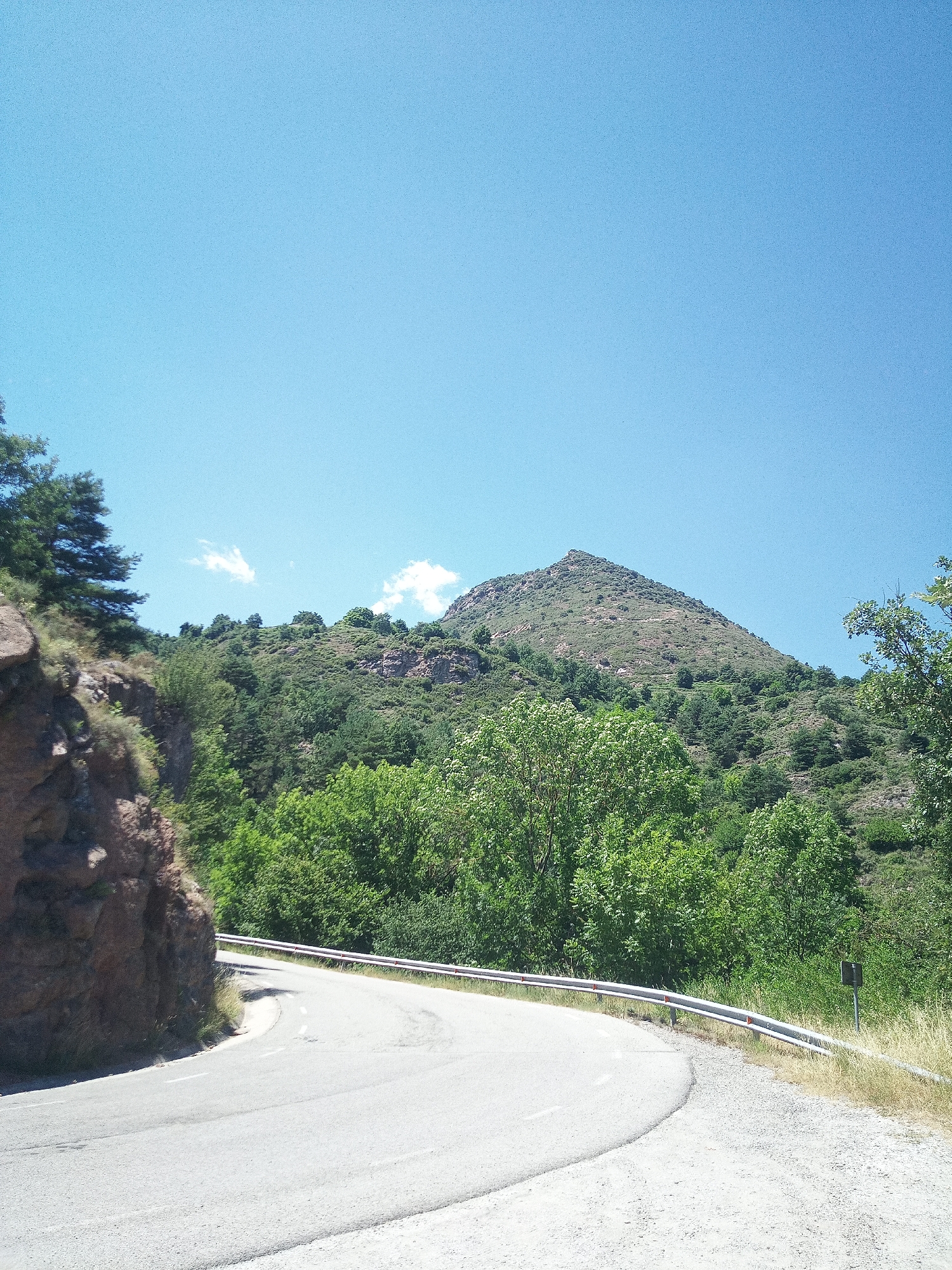 The image depicts a winding road curving through a mountainous, forested landscape under a clear blue sky. The road is paved and has white lane markings. A metal guardrail runs along the edge of the road, separating it from the steep, rocky incline on the left and the dense vegetation on the right. The vegetation consists of a variety of trees and shrubs, with a prominent peak of a mountain dominating the background. There are a few scattered white clouds in the sky. The scene appears to be located in a natural, undeveloped area, likely a mountain pass or a rural route. There are no people or animals visible in the image. The lighting suggests it is daytime, possibly mid-morning or afternoon, given the bright sunlight. The location is identified as being in the city of Berga, Spain, which is known for its mountainous terrain and natural beauty.