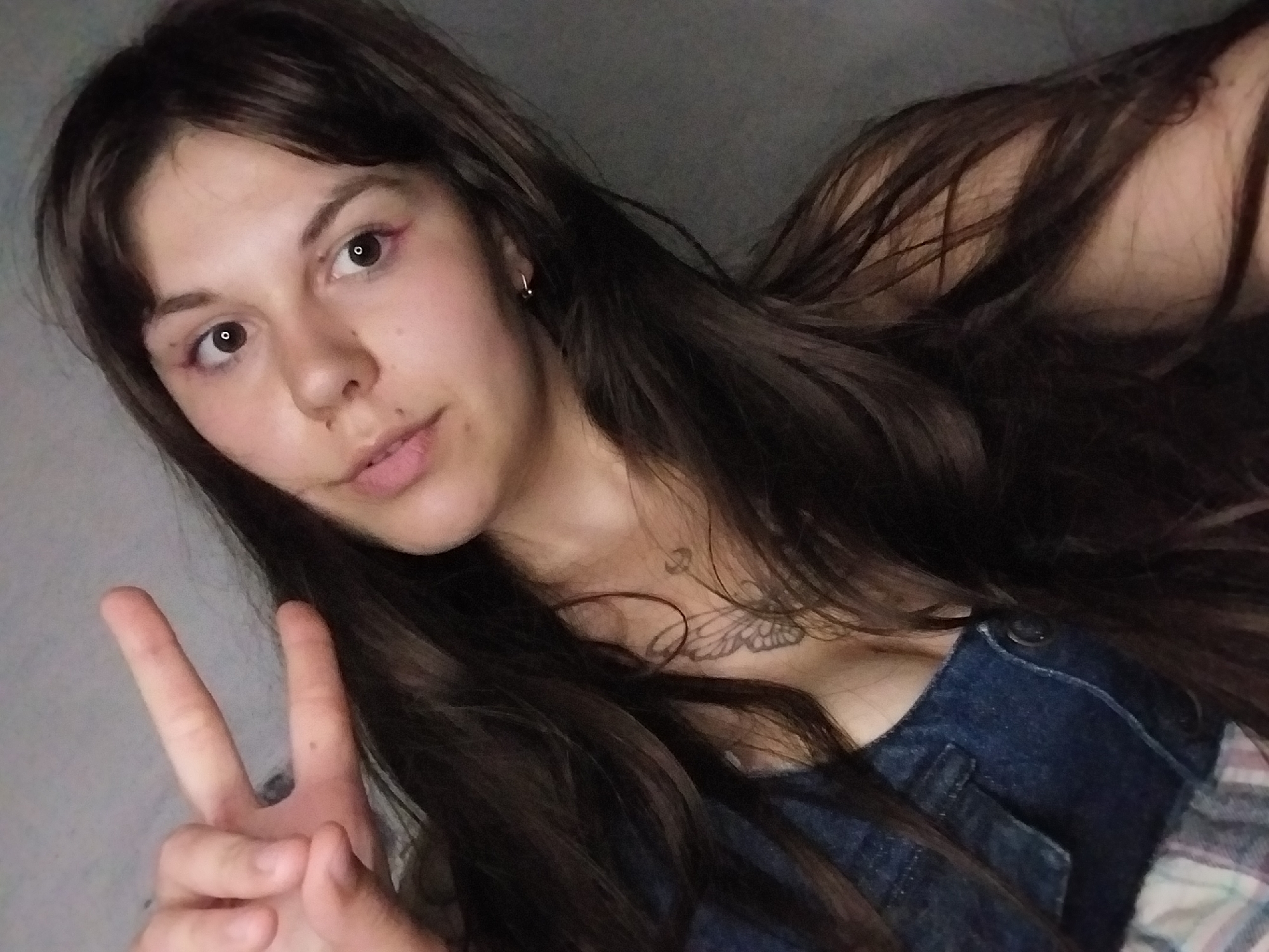 A young woman with long, dark brown hair is taking a selfie, holding up two fingers in a peace sign. She is wearing a denim top, and a tattoo of a butterfly is visible on her neck. Her eyes are wide and looking directly at the camera. The background is a plain, gray wall, and a portion of a plaid blanket is visible at the bottom of the frame. The lighting is soft, and the image appears to be taken indoors.