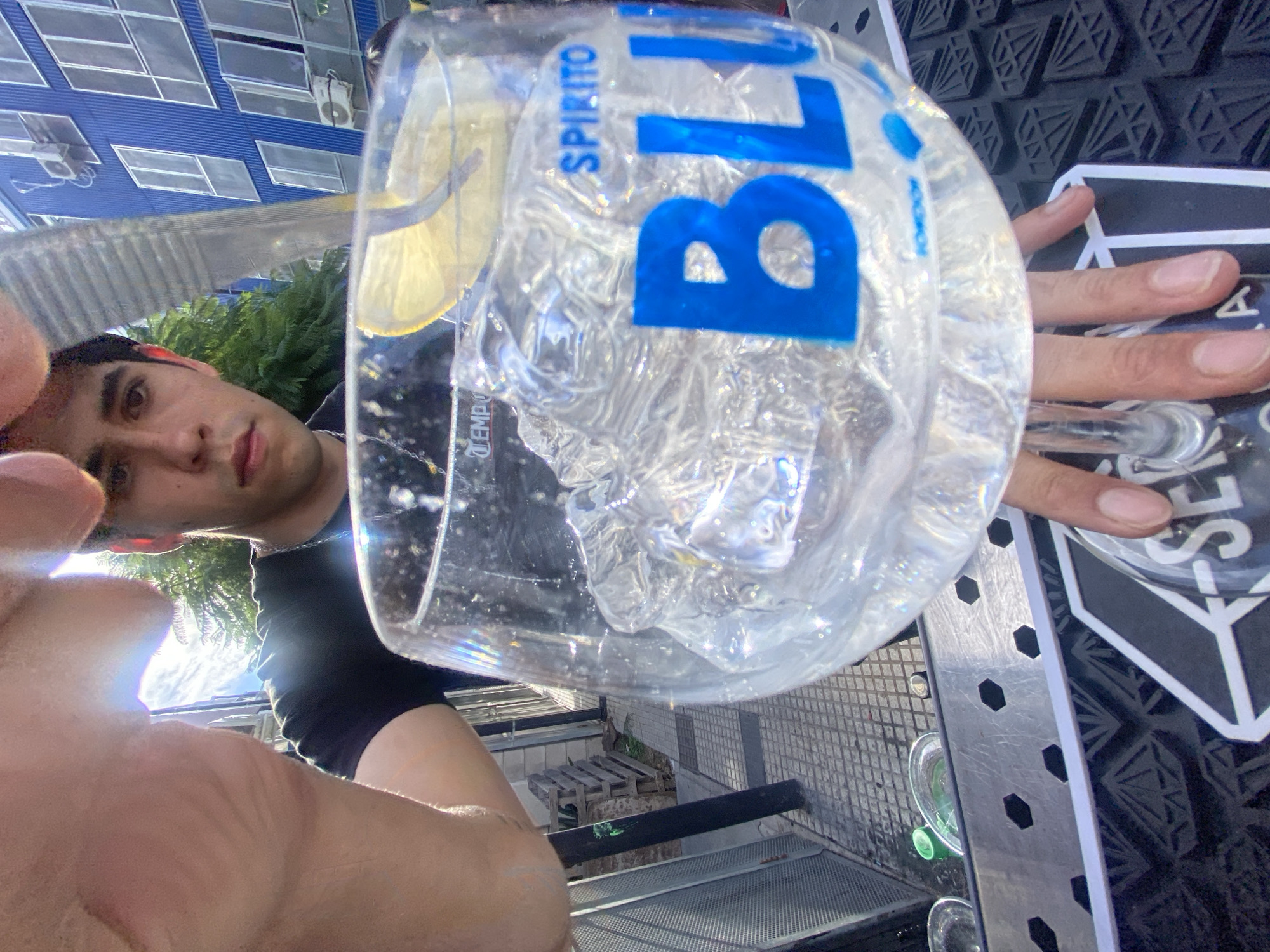 A selfie taken from a low angle shows a man holding a glass with ice and a lemon slice. The glass has the word "BLU" in large blue letters and "SPIRITO" in smaller letters above it. He is standing outside with a view of an urban environment including a blue building with windows. The foreground includes part of his face and hand holding the glass, and the background shows outdoor features suggesting a terrace or patio setting. The location is likely in Buenos Aires, Argentina based on the brand name printed on the glass being local.
