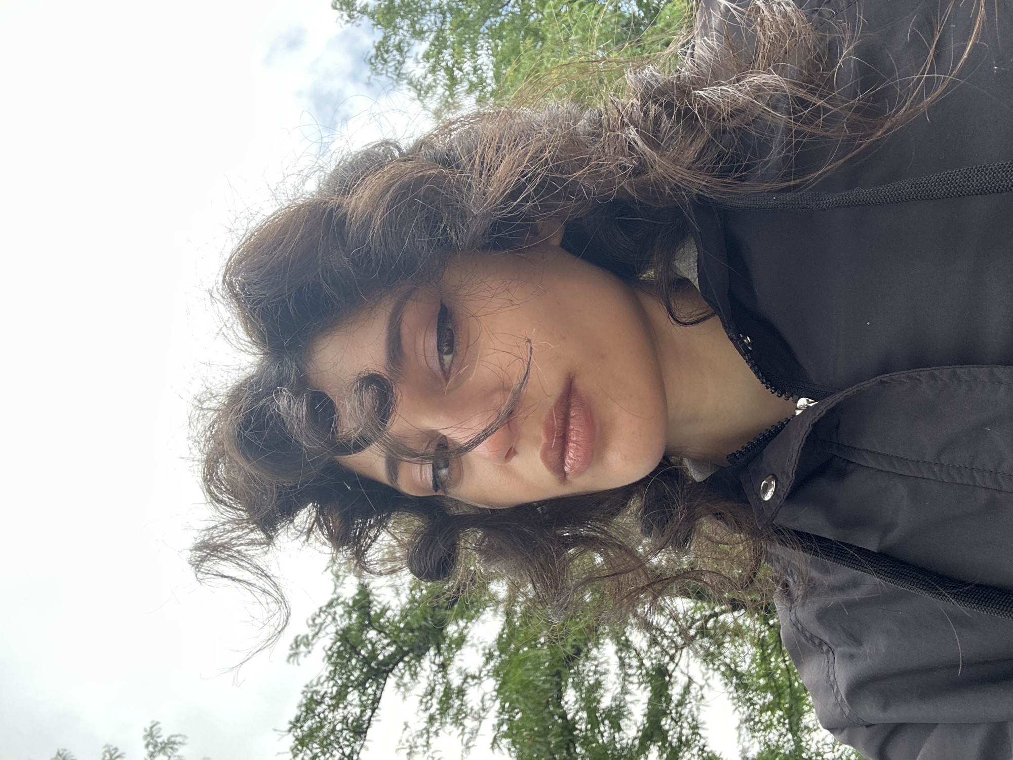 A low-angle, medium shot captures a young woman from the chest up, standing outdoors. She has dark, curly hair that is windblown around her face and shoulders. Her eyes are dark, and she is looking towards the camera. She is wearing a black zipped jacket. In the background, green trees and a cloudy sky are visible. The lighting suggests it is daytime. There is no indication of the location in Sofia, Bulgaria.