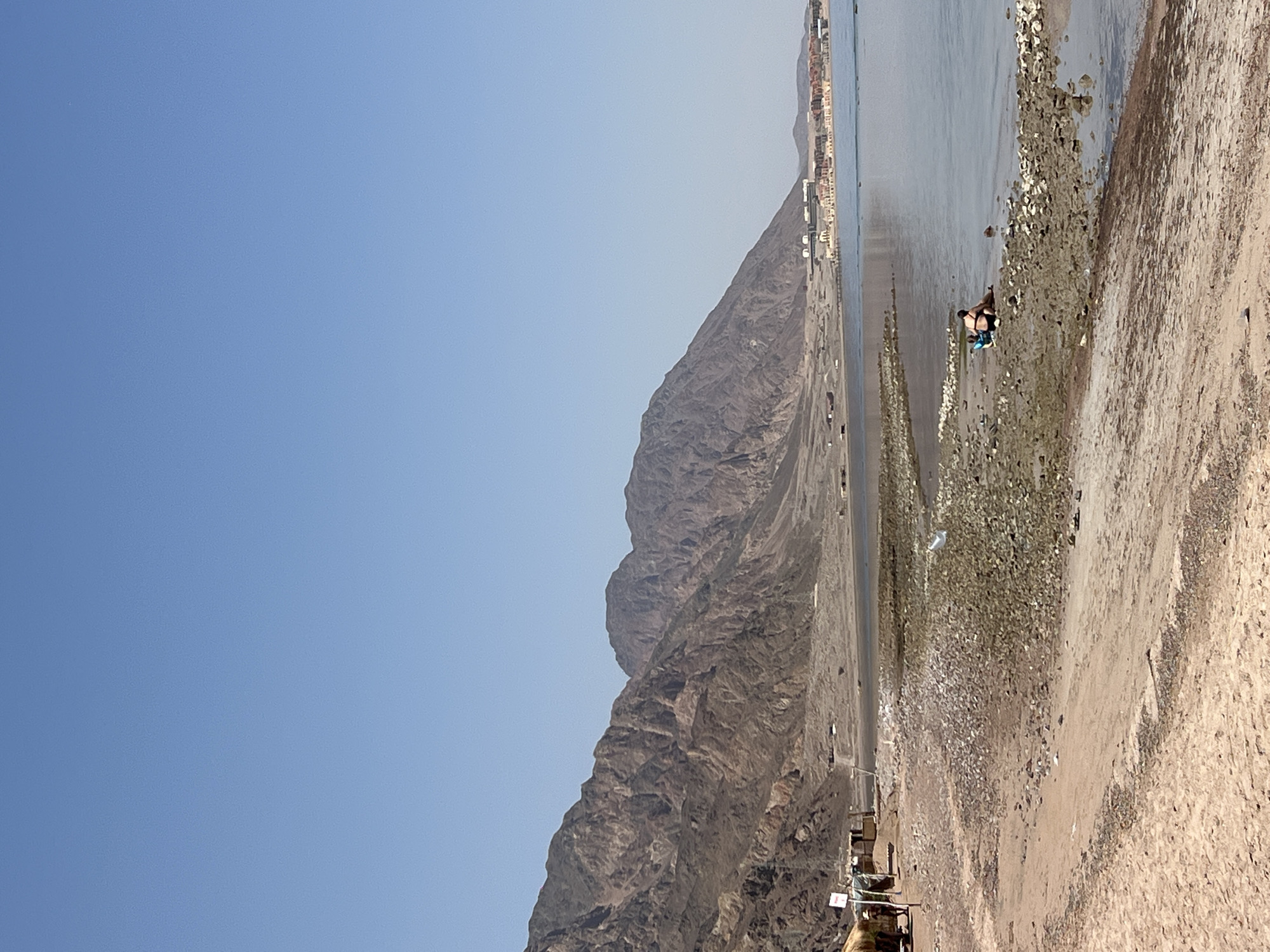 This is a wide-angle, eye-level, outdoor, daytime photograph of a rocky beach and shoreline in a desert mountainous region. The sky is a clear, light blue, taking up the top half of the image. The middle ground features a calm, light blue body of water with a shoreline composed of sand and scattered rocks and pebbles. In the far distance, along the water's edge, are several buildings with terracotta roofs, appearing to be part of a resort or town. The background is dominated by rugged, brown, and tan mountains with visible geological strata.
In the foreground, the beach transitions from sand to a denser area of rocks and gravel. Two individuals are present on the rocky shoreline near the water's edge. One person, wearing black swimwear, is sitting down, while another, also in dark swimwear, is lying down beside them. A blue bag is visible next to the sitting person. Further left on the beach, several small, rustic structures, possibly temporary shelters or market stalls, are visible, constructed from natural materials like straw and wood. A person wearing a hat stands near one of these structures. The overall scene conveys a sense of a quiet, sun-drenched coastal environment in a remote desert area.