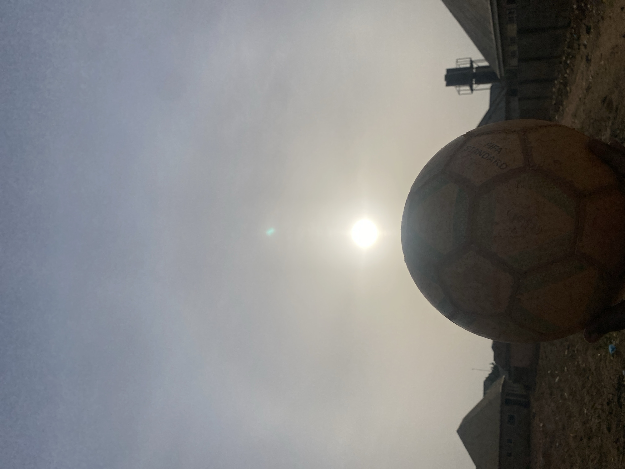 The image, oriented correctly (by rotating it 90 degrees clockwise), presents a close-up perspective of a football against a bright, hazy sky and a distant, subdued landscape.
A well-used football, possibly light brown or tan, occupies the right side of the frame, with its top-right portion extending out of view. The distinct hexagonal and pentagonal panels of the ball are visible, and on one of the panels, the words "FIFA STANDARD" are clearly printed.
The sky dominates the central and left areas of the image, appearing significantly bright and hazy, almost white or light grey, suggesting strong atmospheric diffusion or a high sun. A brilliant sun is prominently positioned in the middle-right of the sky, acting as a strong light source and causing a noticeable lens flare. A small, greenish-blue flare can also be observed to the left of the sun.
In the background, beneath the hazy sky and behind the lower part of the football, dark, silhouetted structures are visible. These appear to be industrial buildings or parts of larger structures, with one exhibiting a multi-tiered or ladder-like element. The ground beneath these structures and to the left of the football is uneven and appears to be dusty, rough terrain or dirt.
The overall lighting suggests daytime, with the sun casting a strong, somewhat obscured glow through the hazy atmosphere. The scene evokes an outdoor setting, possibly an informal playing area or an undeveloped lot, under bright but diffused sunlight.
