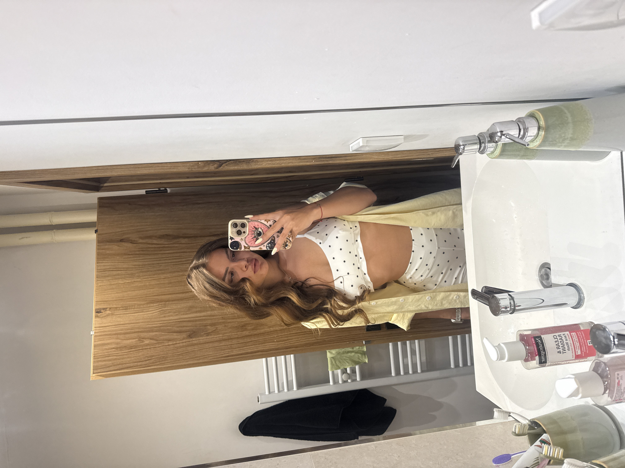 A woman is taking a mirror selfie in a bathroom. She is wearing a white polka dot crop top and a yellow shirt. She has long, wavy brown hair and is wearing several rings on her fingers. In her hand, she is holding a phone with a pink phone case decorated with a pink eye and eyelashes. The bathroom has a wooden vanity, a white sink with chrome faucets, and a white towel rack with a black towel hanging on it. A bottle of Clearasil Rapid Facial Wash is visible on the sink. The lighting suggests it is daytime. The woman appears to be in a modern bathroom.