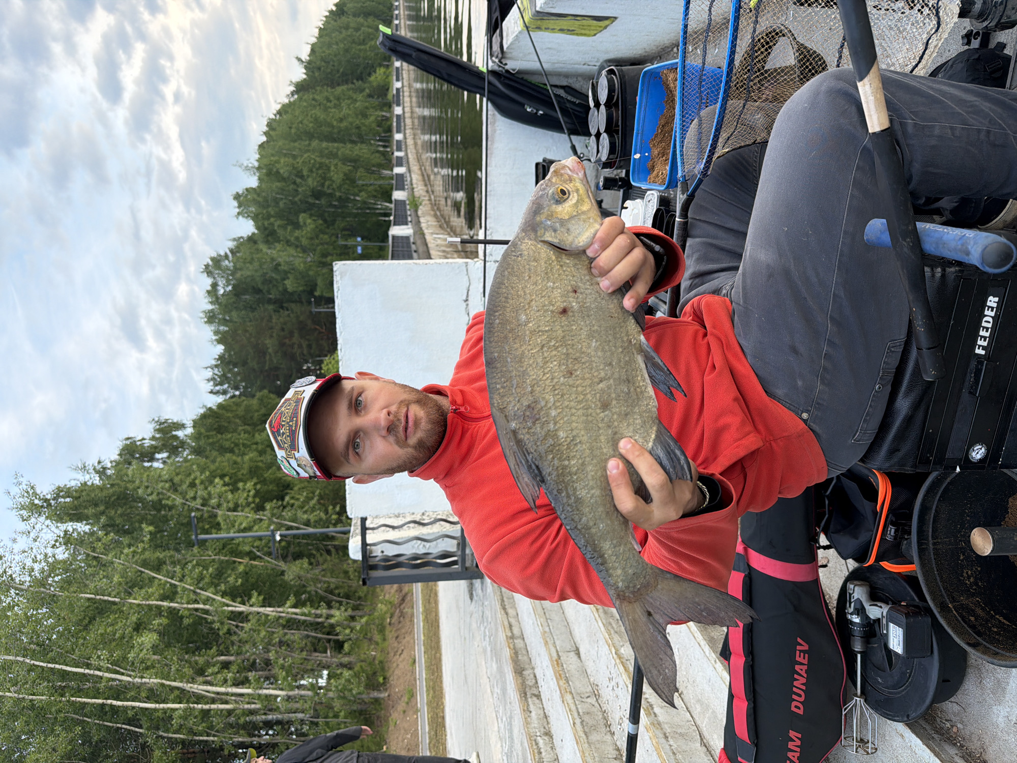 A man is depicted seated outdoors, holding up a freshly caught, large fish, likely a bream. He wears a red fleece jacket, dark trousers, and a multi-badged baseball cap, looking directly at the camera with a neutral expression.
The setting is an embankment or pier, possibly made of concrete, beside a body of water, which appears to be a river or canal. In the background, across the water, a dense treeline and some distant structures are visible. The sky is overcast with clouds.
Around the man, various fishing gear is arranged. A blue tray containing groundbait or bait is placed near a fishing net. A black bag or box labeled "FEEDER" is next to him, hinting at the type of fishing. Another black and red bag with "TEAM DUNAEV" printed on it is also visible, along with a cordless drill fitted with a mixing whisk, commonly used for preparing groundbait. Parts of fishing rods can be seen resting in holders.
The image captures an angler during a fishing outing in Chaikovsky, Russia, highlighting his catch and specialized equipment. The visible text "FEEDER" and "TEAM DUNAEV" point to specific brands or styles of fishing.