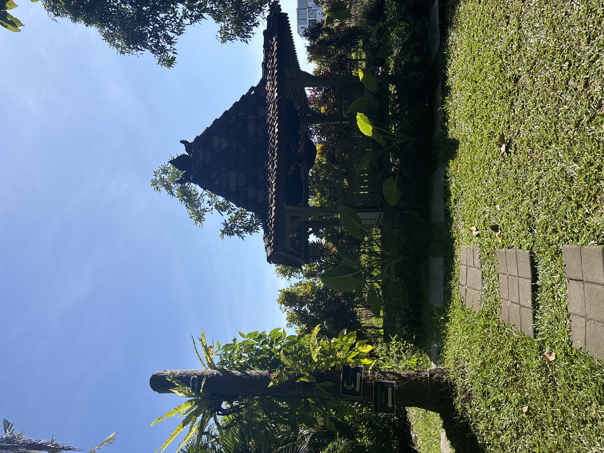 The image depicts a serene garden scene in Mengwi, Indonesia. In the foreground, a lush green lawn is partially covered by stepping stones, forming a pathway. To the left, a tree trunk serves as a support for various plants, including ferns and a bird's nest fern. Two signs are attached to the tree trunk, one with a circular symbol and text, and another labeled "MAHAMUSTIKA SHALA" with an arrow.
Dominating the mid-ground is a traditional Balinese-style gazebo with a dark, tiled roof supported by wooden pillars. The gazebo is surrounded by dense foliage, including large-leafed plants. In the background, more trees and shrubs create a vibrant green backdrop, with hints of red and yellow foliage. To the far right, a modern building with white walls and windows is visible above the trees. The sky is clear and bright blue, indicating a sunny day.