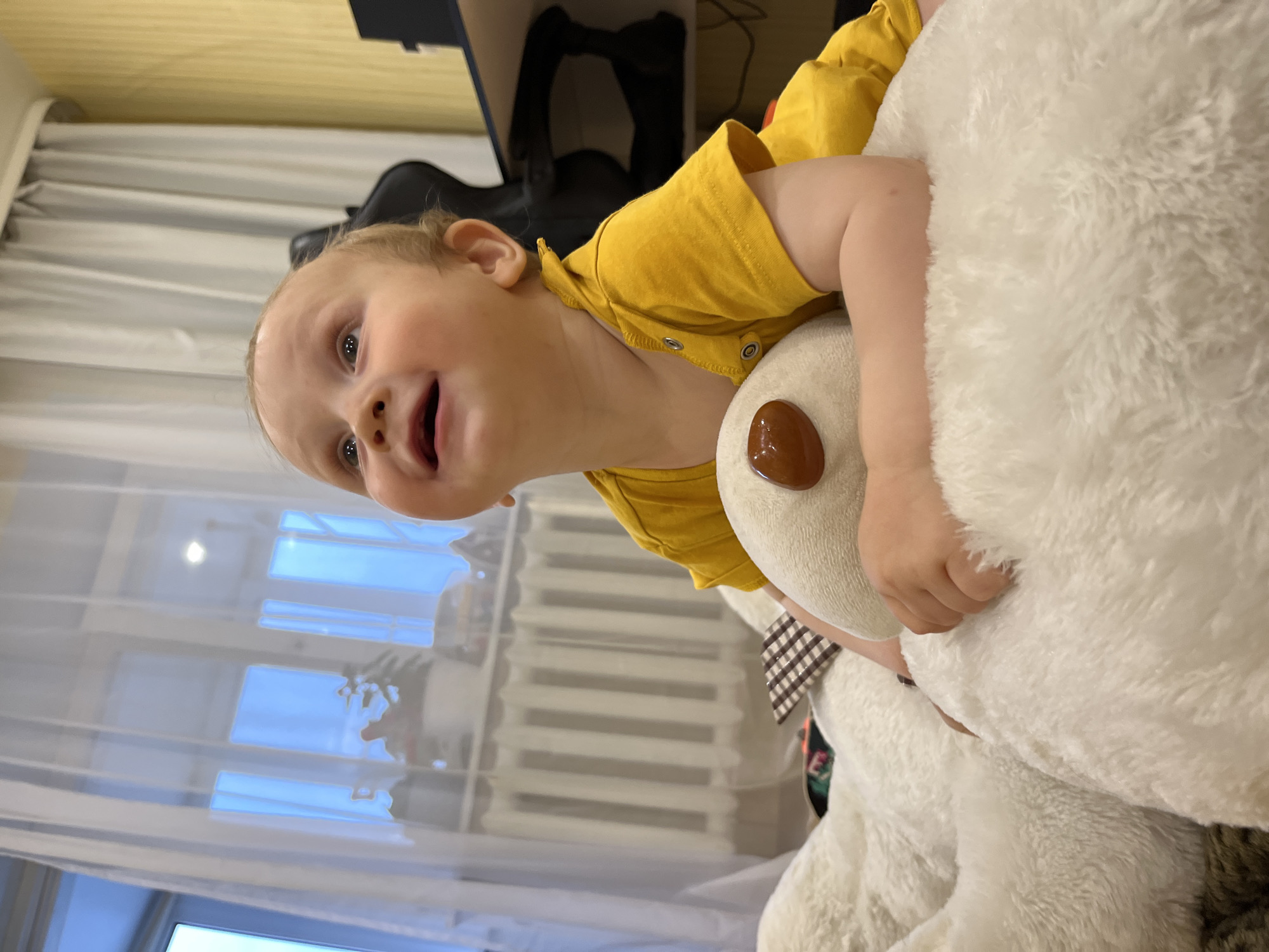 A light-skinned infant, estimated to be a baby or young toddler, is depicted indoors in the city of Chaikovsky, Russia. The infant is positioned on its stomach, supported by its forearms, which rest upon a large, white, shaggy textile object. The infant wears a yellow short-sleeved, collared top secured with snap fasteners and exhibits a smiling expression, with eyes directed upwards and slightly to the left. The infant's hands are placed on the white textile and are grasping a smaller, beige plush toy that features a single, glossy, brown, circular element.
The background on the left side of the frame displays a window covered by sheer white curtains, through which a bright blue outdoor light is visible. A white radiator is situated beneath the window. The right side of the background reveals a textured, light yellow wall and a partial view of a dark-colored office chair positioned next to a dark desk. The scene is illuminated by natural light emanating from the window, with minor reflections visible on glossy surfaces.